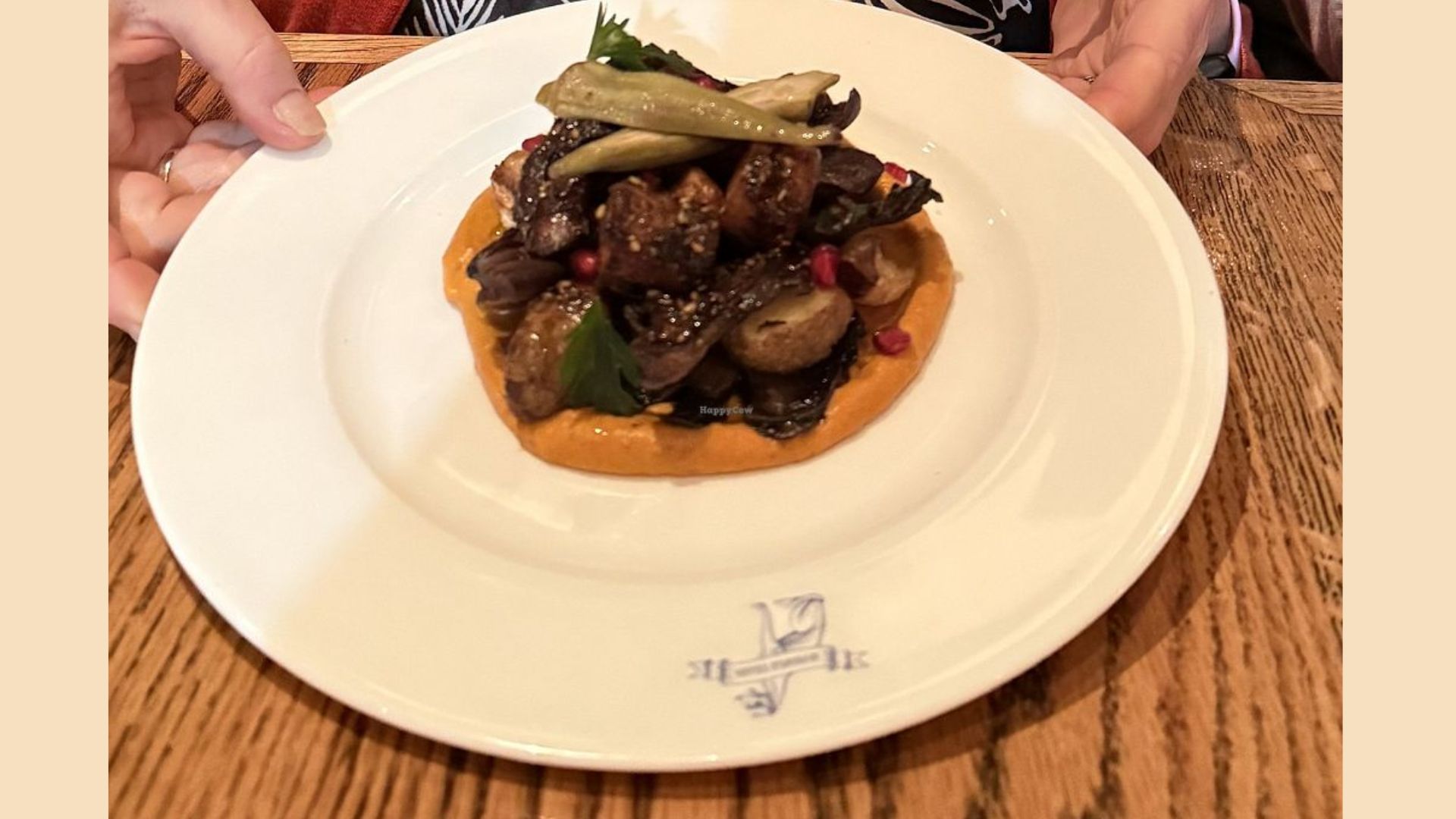 "Plated dish of glazed meat and roasted potatoes over an orange puree, garnished with pomegranate seeds, okra, and herbs, held by two hands over a wooden table"