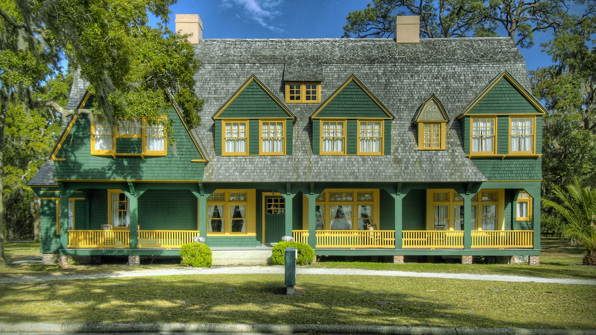 "Front view of a two-story green wooden house with yellow trim and gabled dormer windows, marked AD 1896, surrounded by trees and lawn on a sunny day"