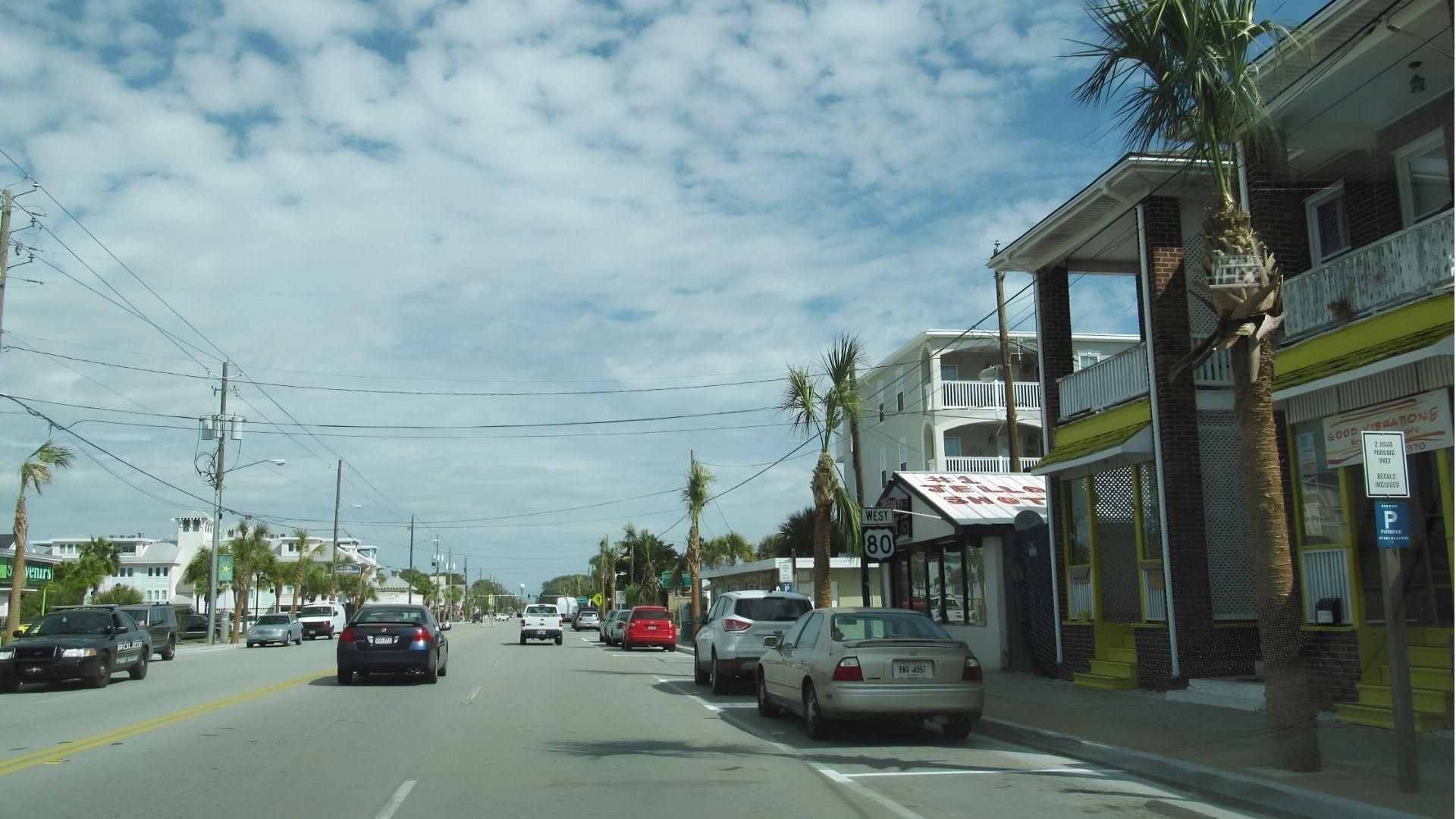 Street-level view of Butler Avenue, the main road through Tybee Island, lined with palm trees, beach shops, and low-rise buildings.