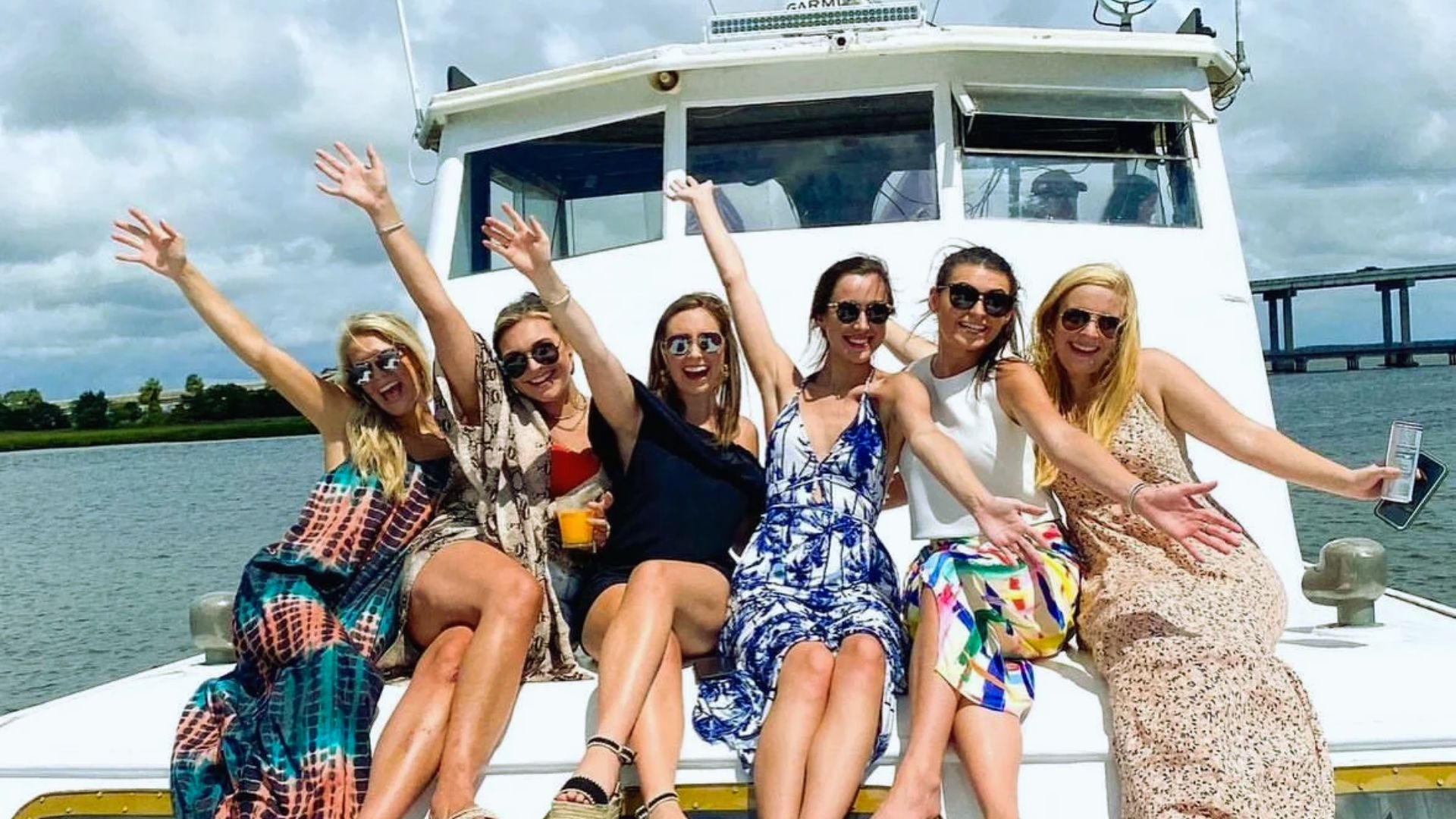 "Six women in sundresses wave and smile while sitting on the bow of a boat on a sunny day."