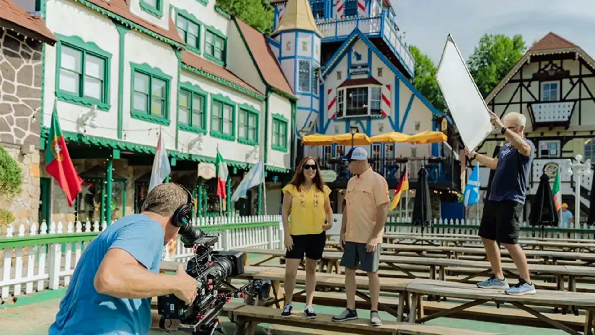 "Film crew records two people on picnic benches in front of Bavarian-style buildings with flags and timber framing."