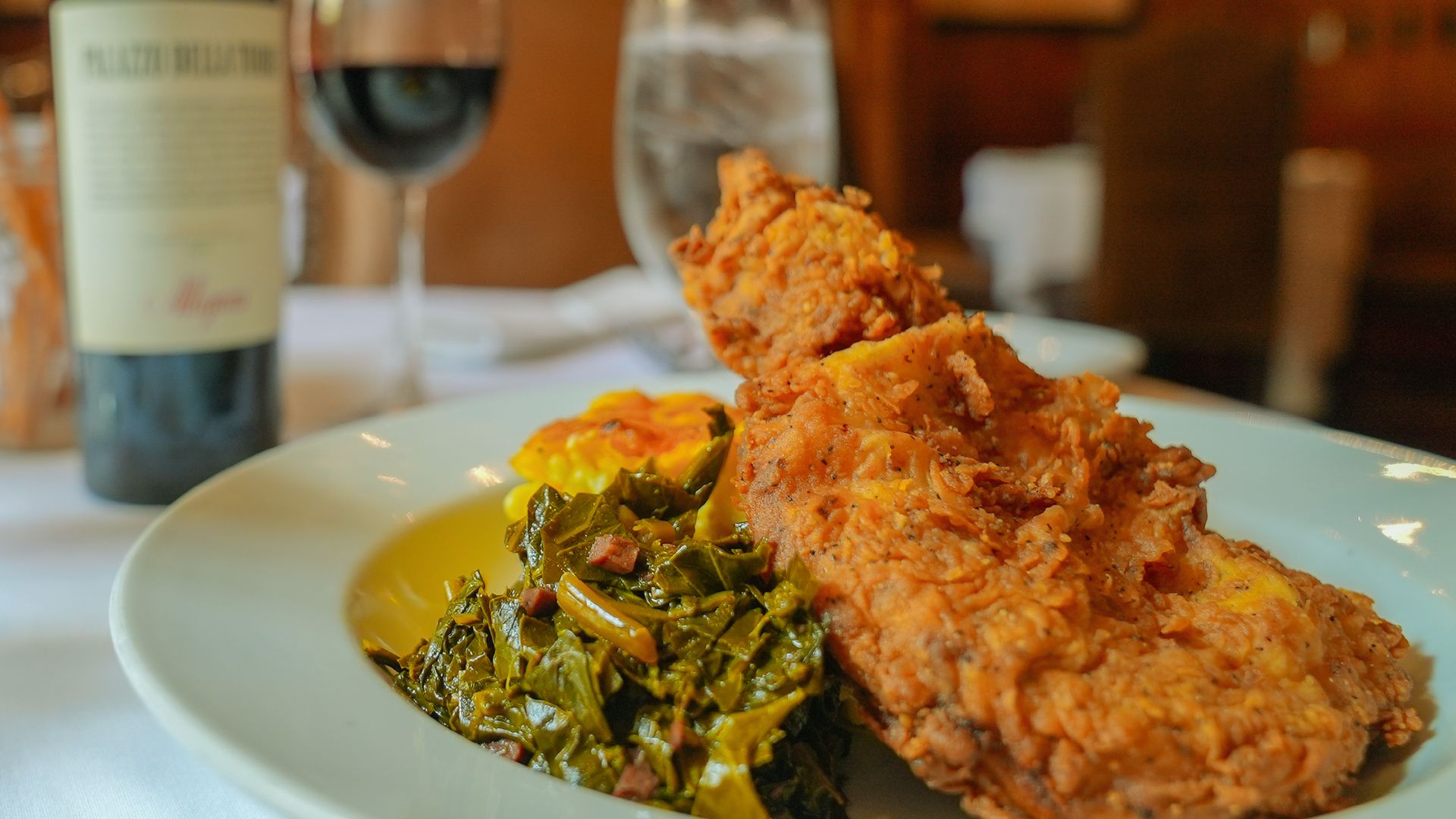 "Plate of golden fried chicken with collard greens and mac and cheese, served with a glass of red wine and a wine bottle in the background"