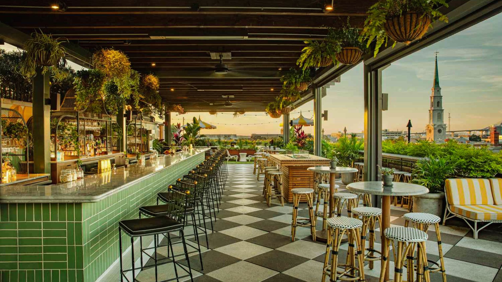 "Rooftop bar at sunset with black and white checkered tile floor, green tiled bar with black stools, hanging plants, bistro tables, and a city skyline with a church steeple in the background"