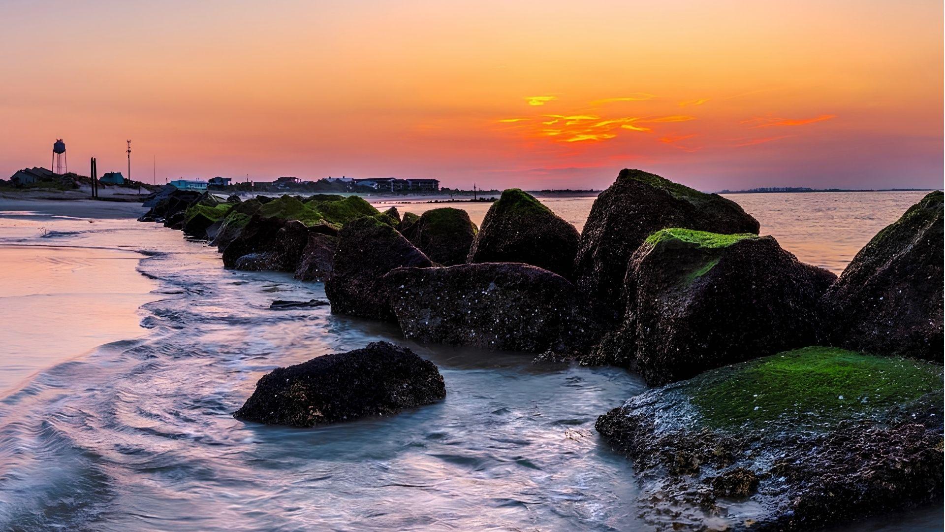 Moss-covered rocks along the shoreline of Tybee Island during a fiery orange sunset, with calm Atlantic water.