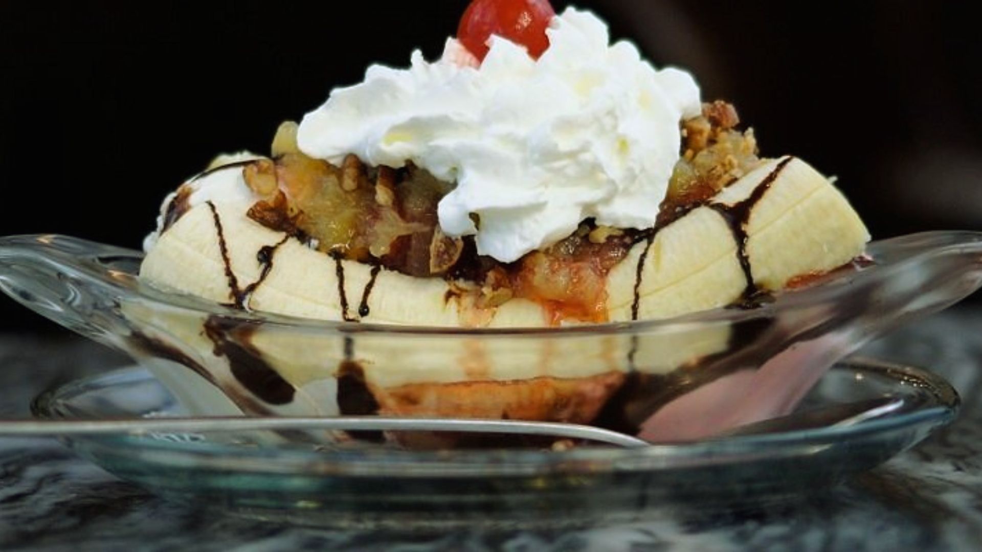 "Banana split dessert in a glass boat dish with a sliced banana, whipped cream, a cherry, chopped nuts, fruit topping, and chocolate drizzle"