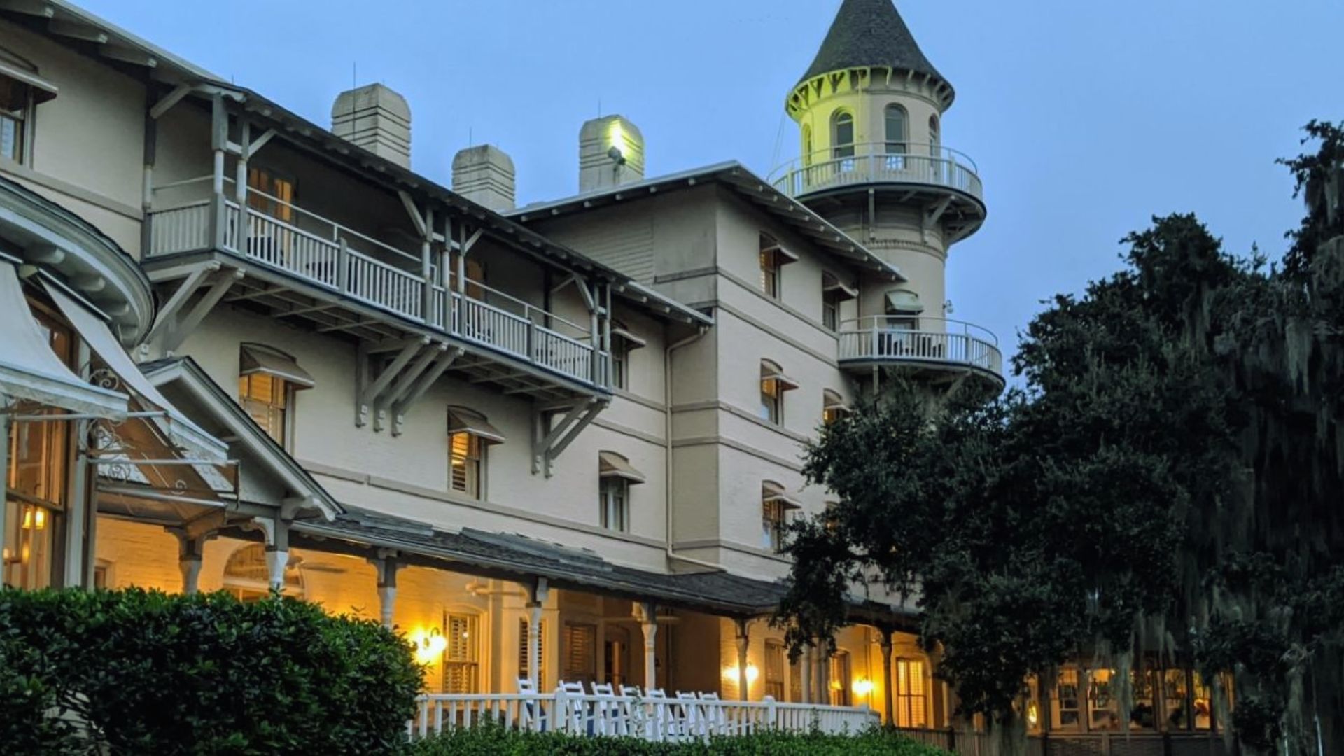 "Exterior of a large historic hotel at dusk with white balconies, warm glowing windows, and a lit turret tower beside trees draped in Spanish moss"