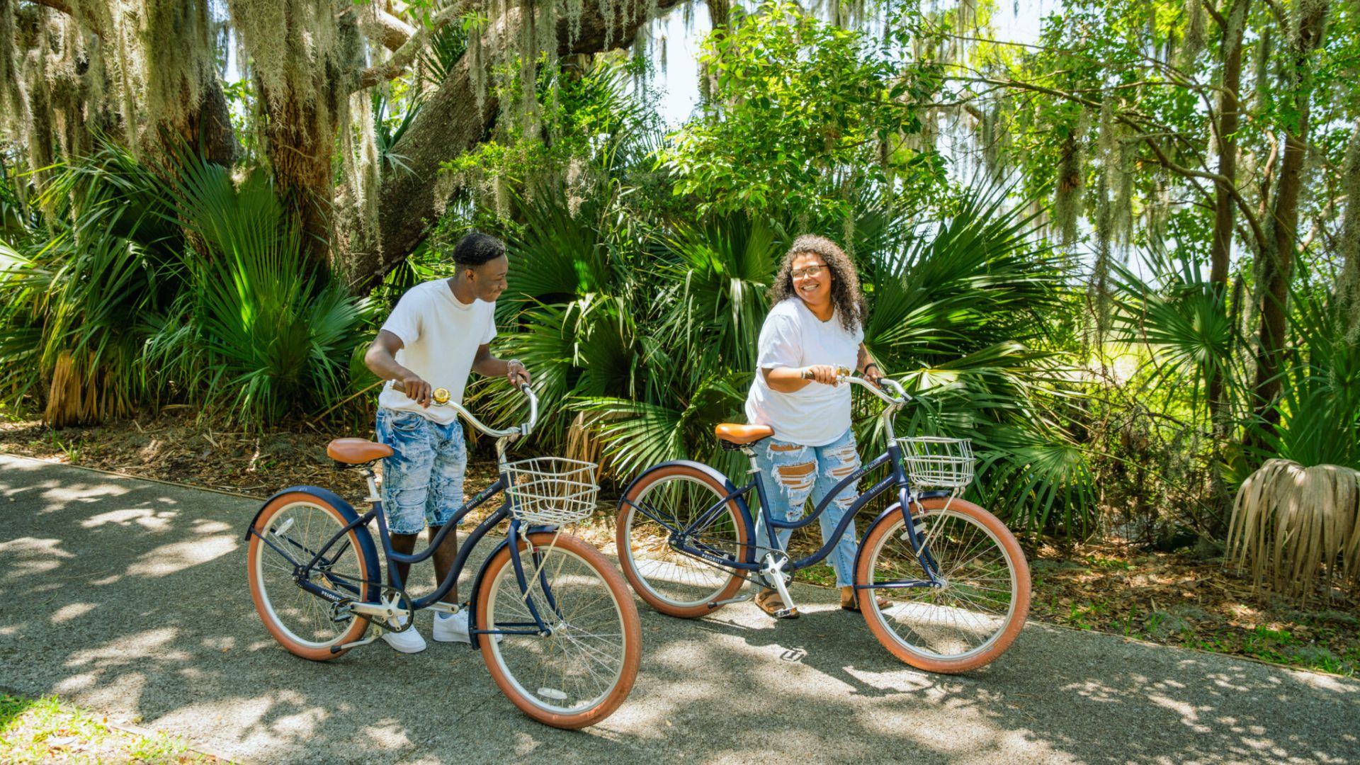 "Two people smiling on navy blue cruiser bikes with white baskets on a shaded paved path surrounded by lush green palms and trees with hanging moss"
