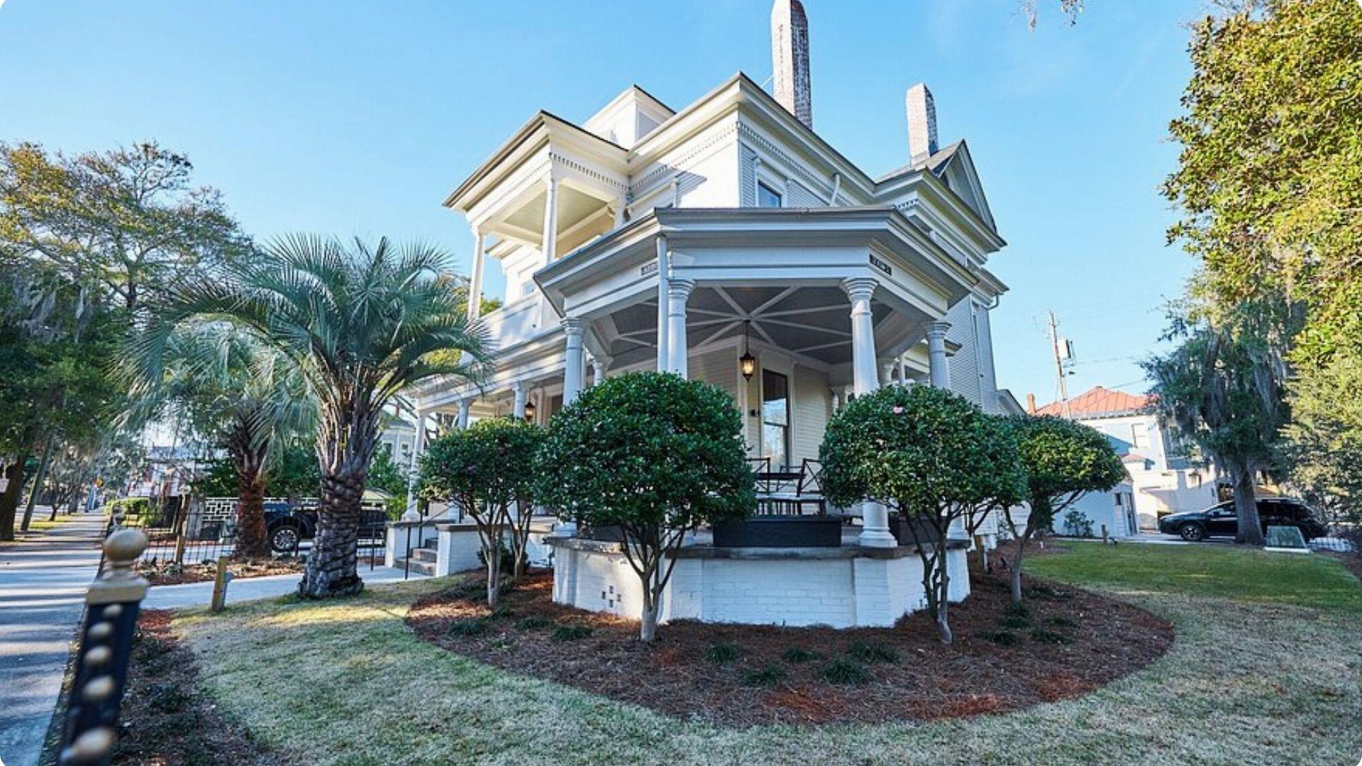 A gracious Victorian-era white house with a wraparound porch, decorative columns, palm trees, and manicured shrubs.