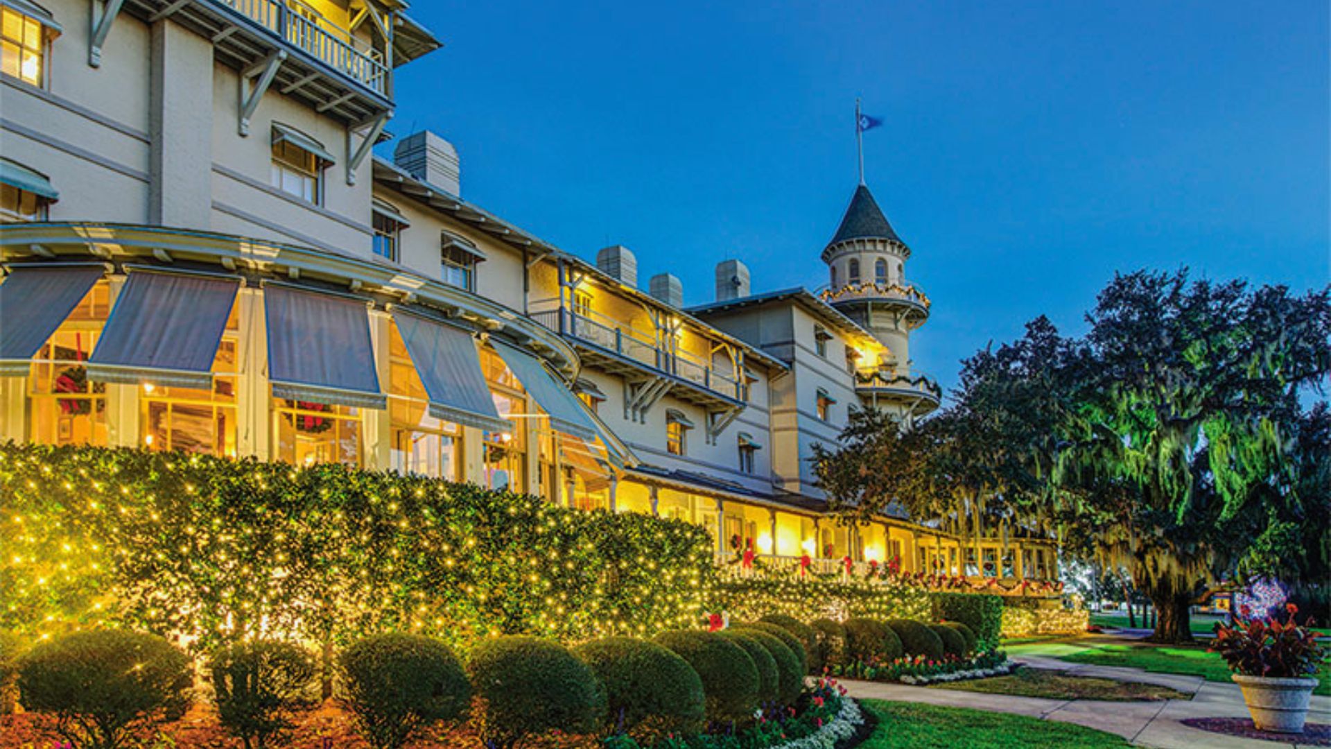 "Grand historic hotel at twilight decorated with string lights on hedges and holiday wreaths, featuring awnings, a turret with a flag, and warm light from windows"