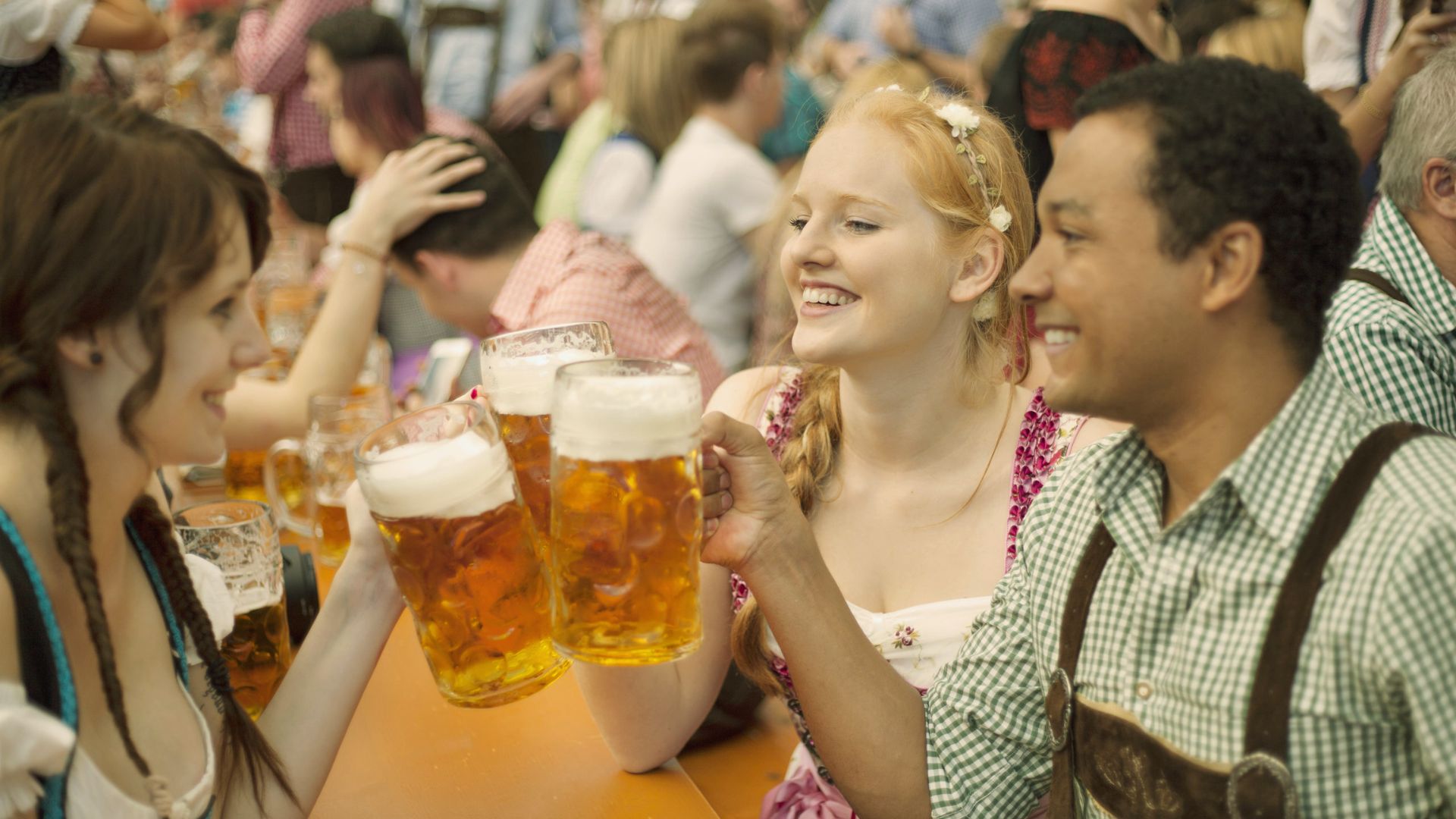 "Three friends in traditional Bavarian dress toast with large beer mugs at a crowded Oktoberfest table."