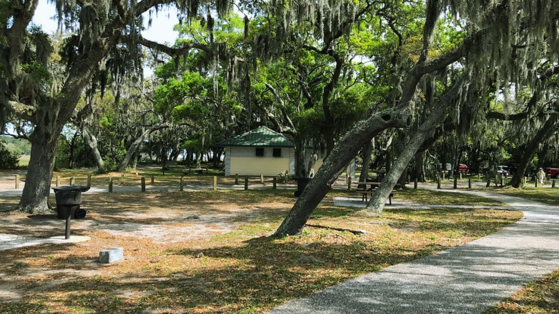 "Shaded park with Spanish moss on oak trees, paved path, picnic table, grill, and small building."