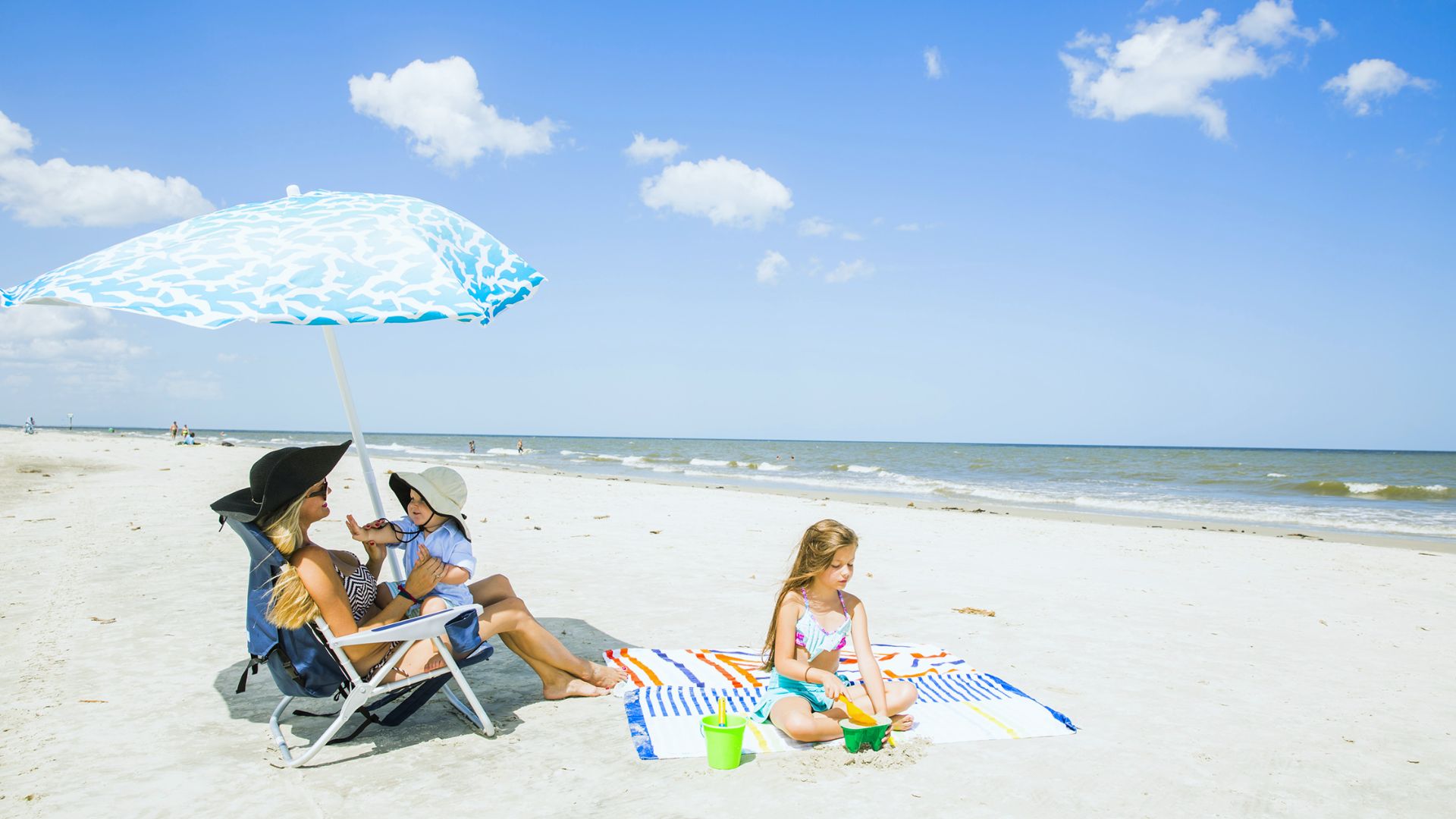 "Woman in a black sun hat sitting in a beach chair under a blue patterned umbrella holding a toddler, while a young girl plays in the sand nearby on a sunny beach"
