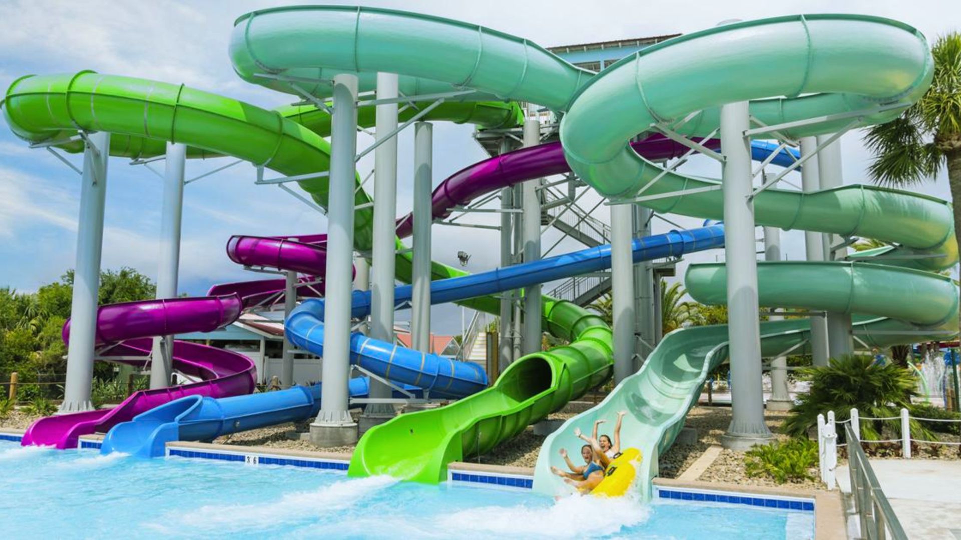 "Colorful water park with green, purple, and blue spiral waterslides emptying into a pool, two people on a yellow raft splashing down the light green slide"
