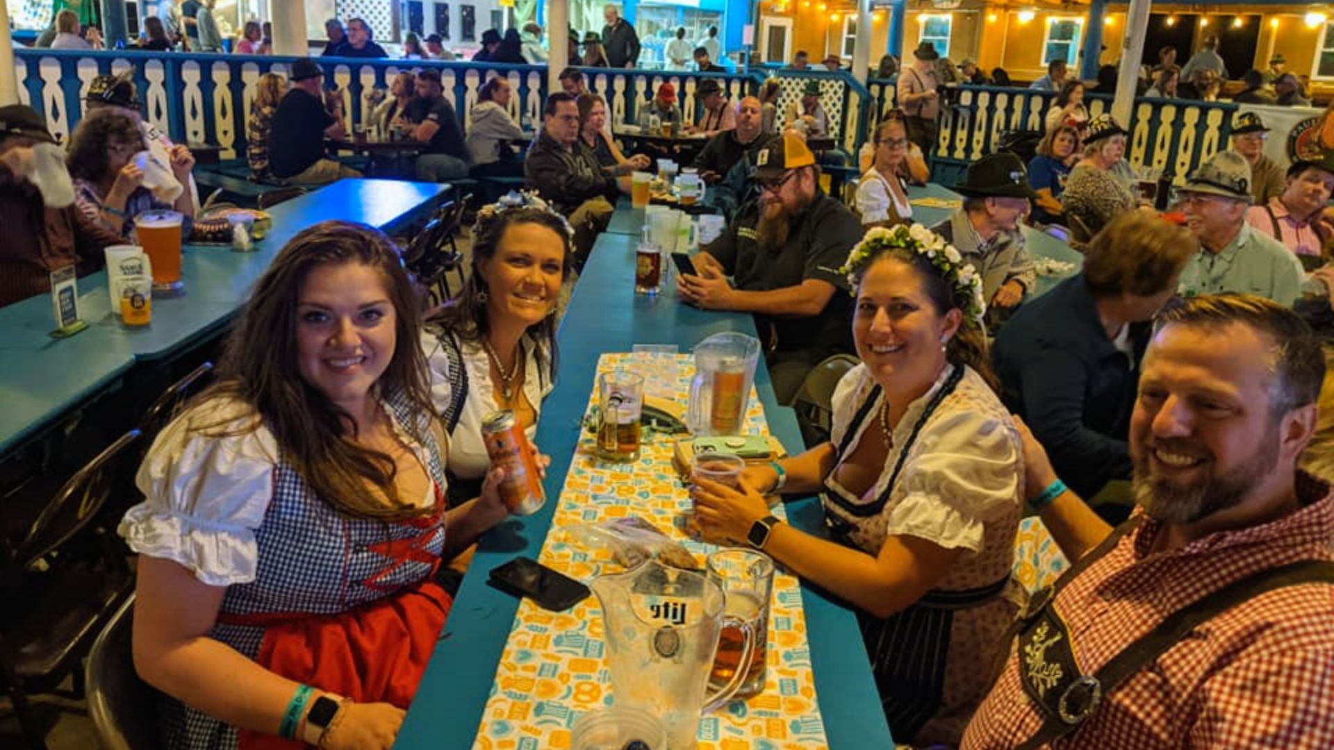 "Group in dirndls and lederhosen smile at a beer hall table with beer mugs and a pitcher."