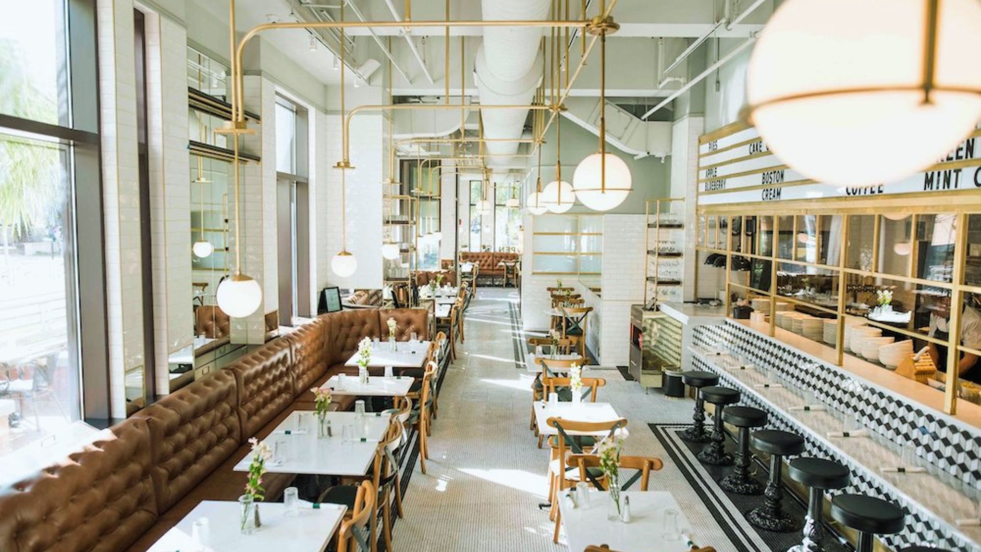 "Bright, modern restaurant interior with brown tufted banquettes, white tables, globe pendant lights with gold fixtures, black and white tile floor, and a menu board listing pies and cakes"