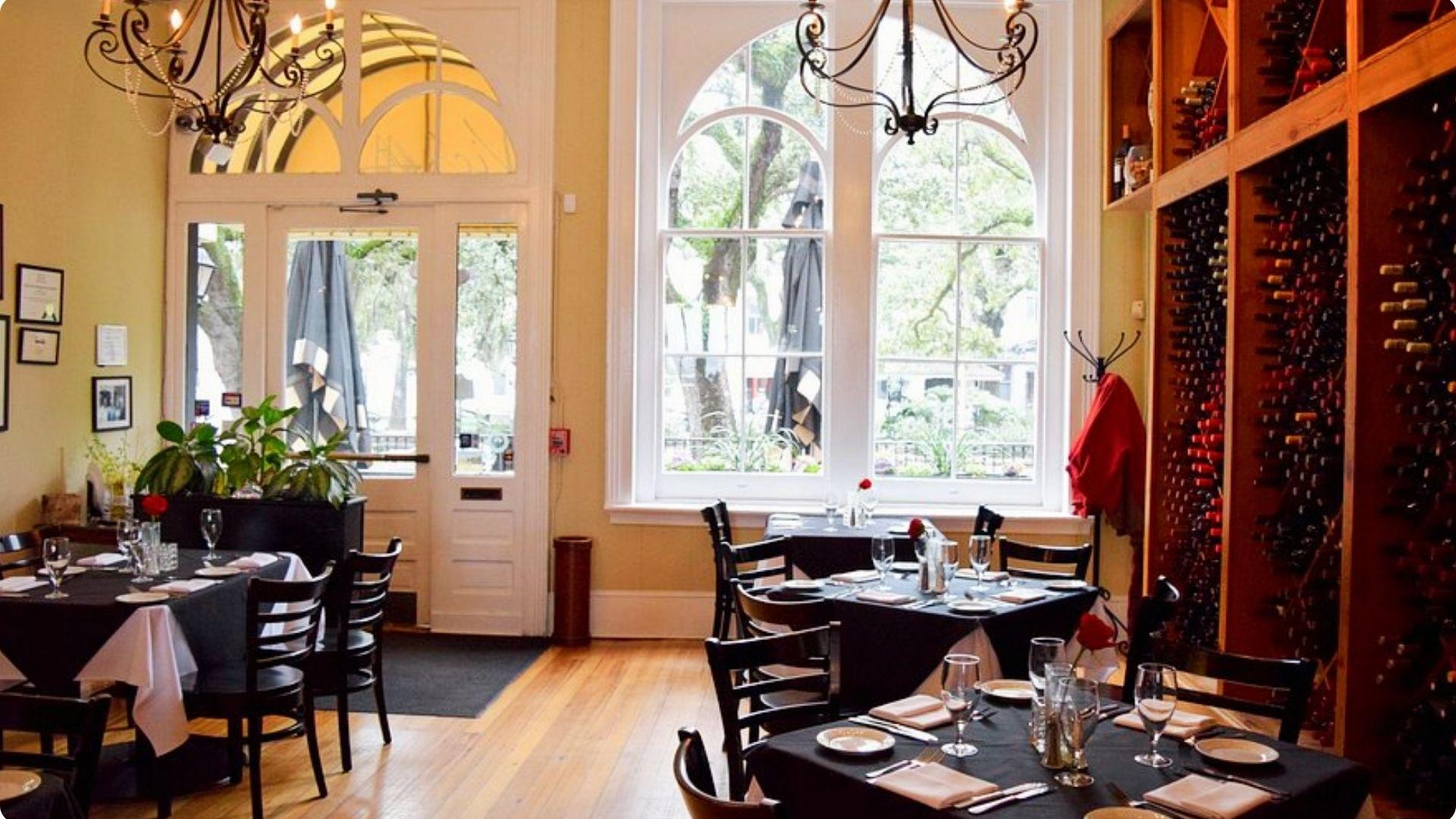 Intimate Italian restaurant dining room with black-draped tables, tall arched windows overlooking a tree-lined square, iron chandeliers, and a large wine rack.