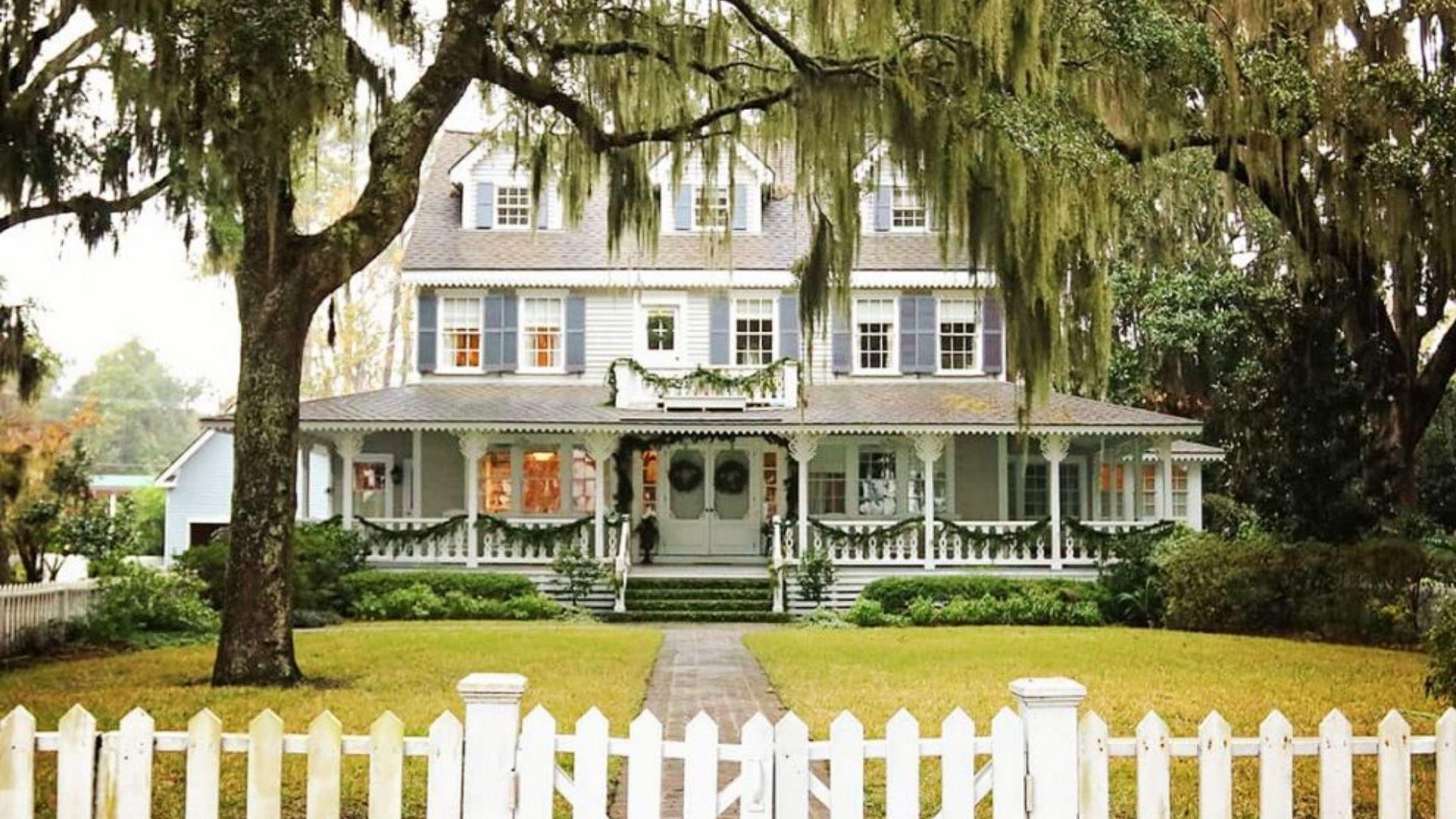 Historic Savannah home with wraparound porch decorated with green garlands and wreaths, framed by Spanish moss trees and white fence