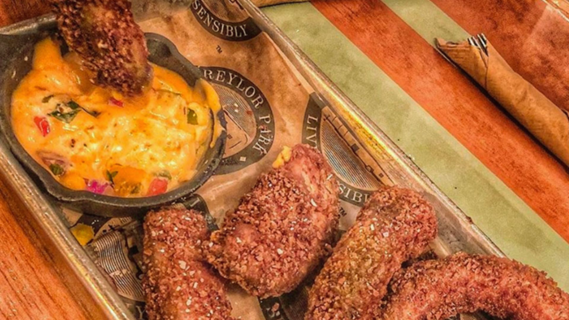 "Metal tray with breaded fried appetizers being dipped into a small cast iron skillet of orange cheese dip with diced peppers, lined with branded newspaper-style paper"