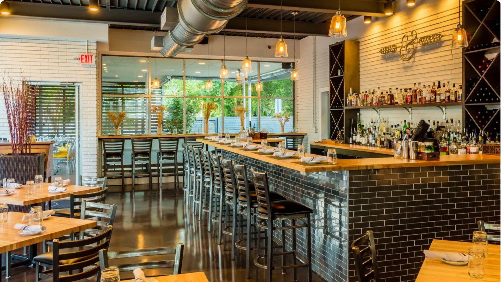 Bright, modern restaurant interior with a long wooden bar topped with dark subway tiles, pendant lighting, white brick walls, and a view of an outdoor patio.