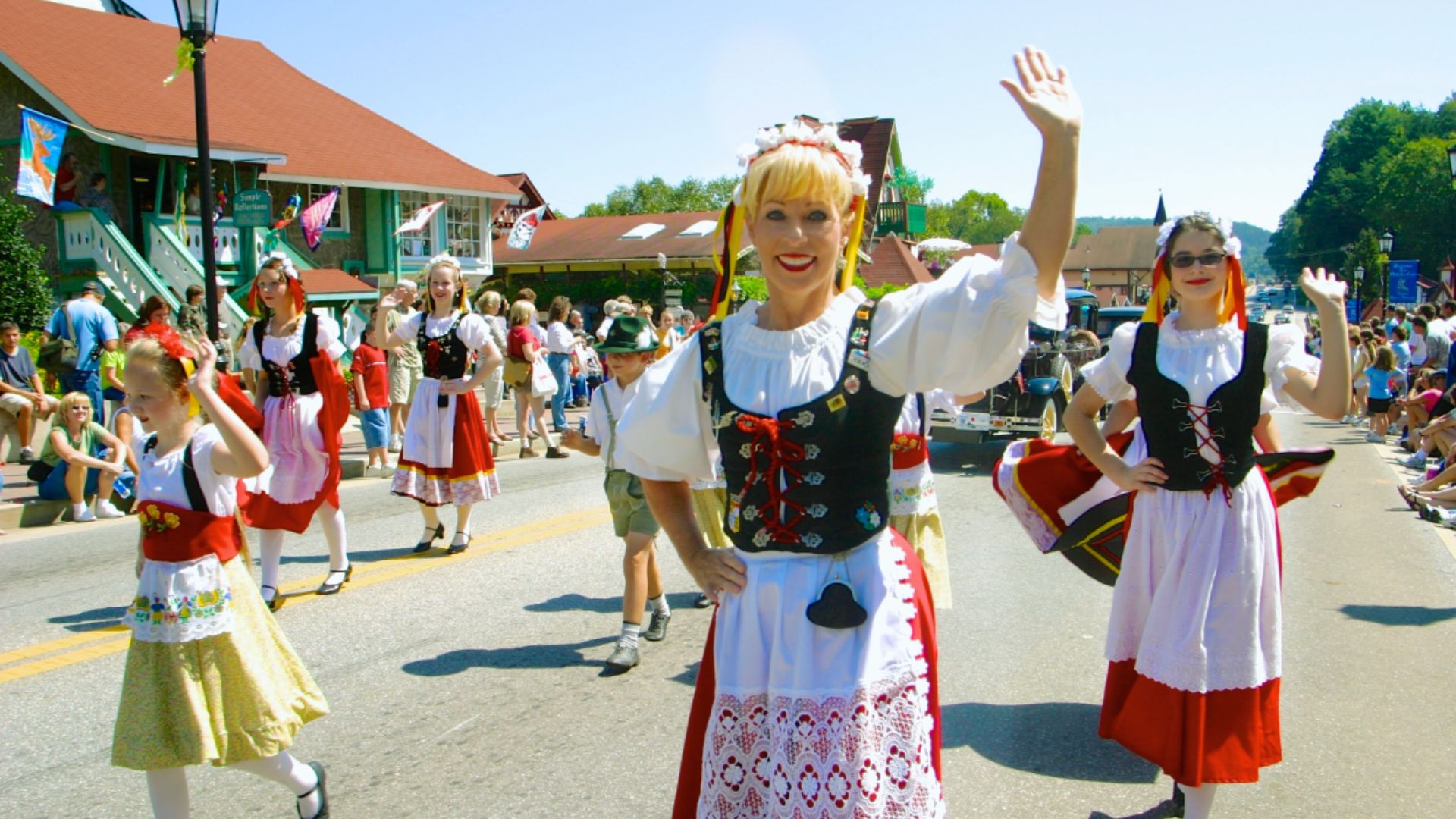 "Women and children in folk costumes wave while walking in a sunny street parade with spectators."