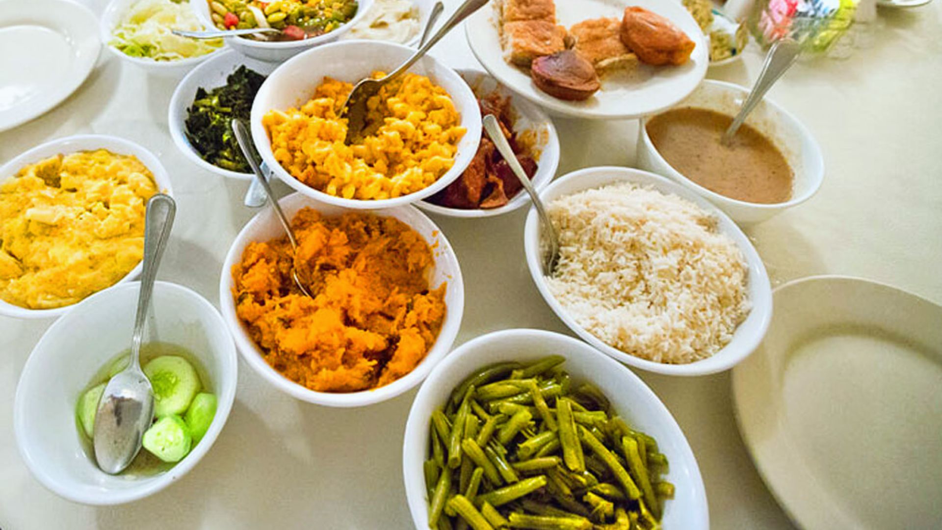 "Overhead shot of a Southern-style spread with multiple white bowls containing mac and cheese, mashed sweet potatoes, green beans, white rice, collard greens, coleslaw, cucumber salad, gravy, and cornbread on a white table"