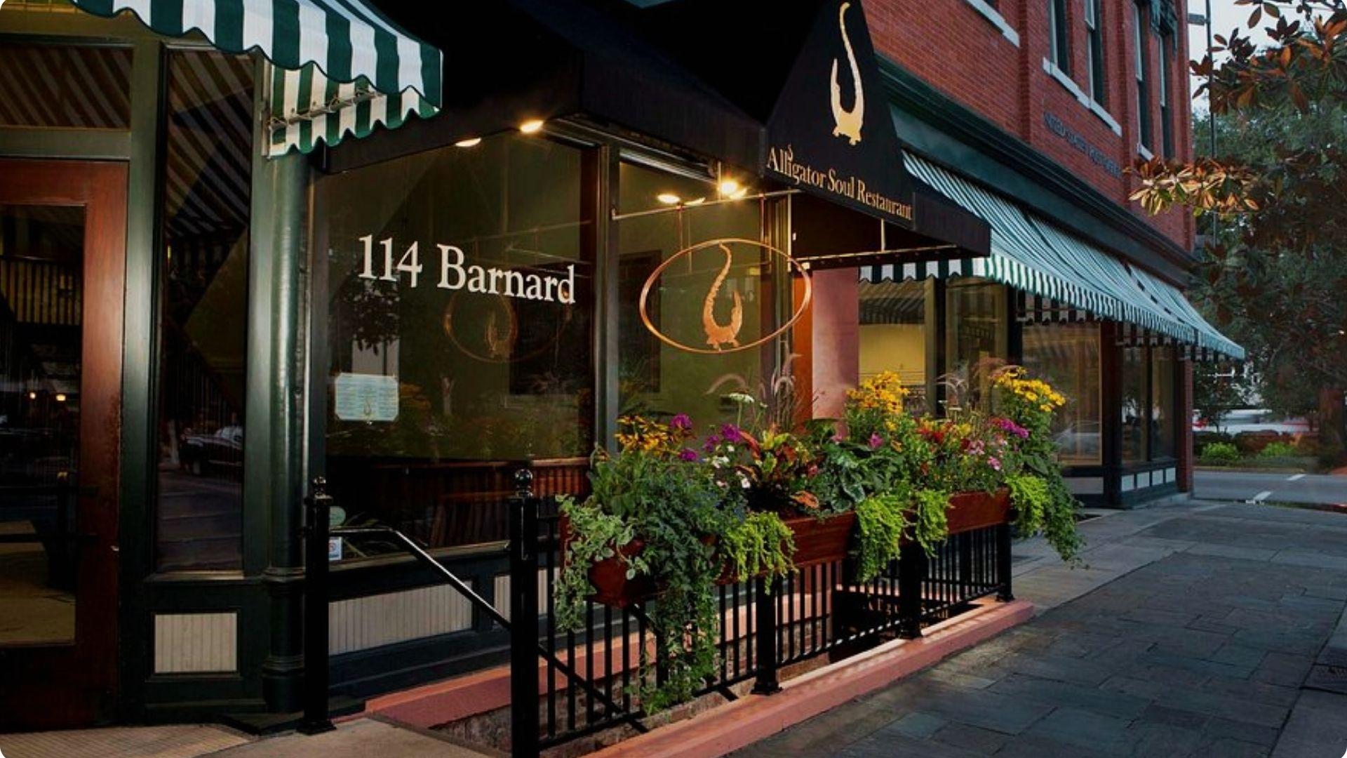 Nighttime exterior of Alligator Soul Restaurant, featuring dark awnings, an alligator logo, and colorful flower boxes along a black iron fence.