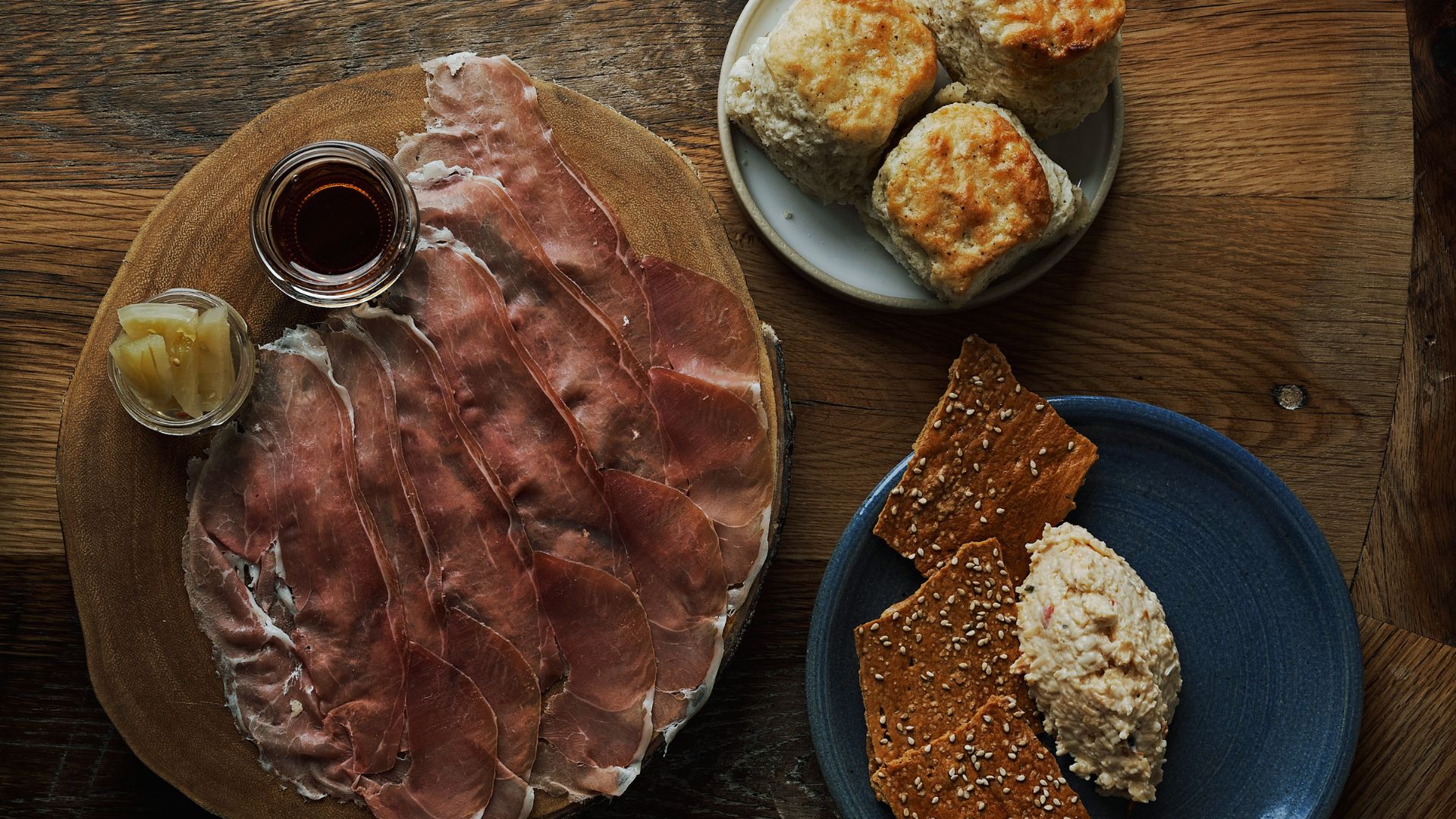 "Overhead view of charcuterie spread on a wooden table with sliced cured meat, small jars of honey and pickles on a wood board, biscuits on a white plate, and sesame crackers with pimento cheese on a blue plate"