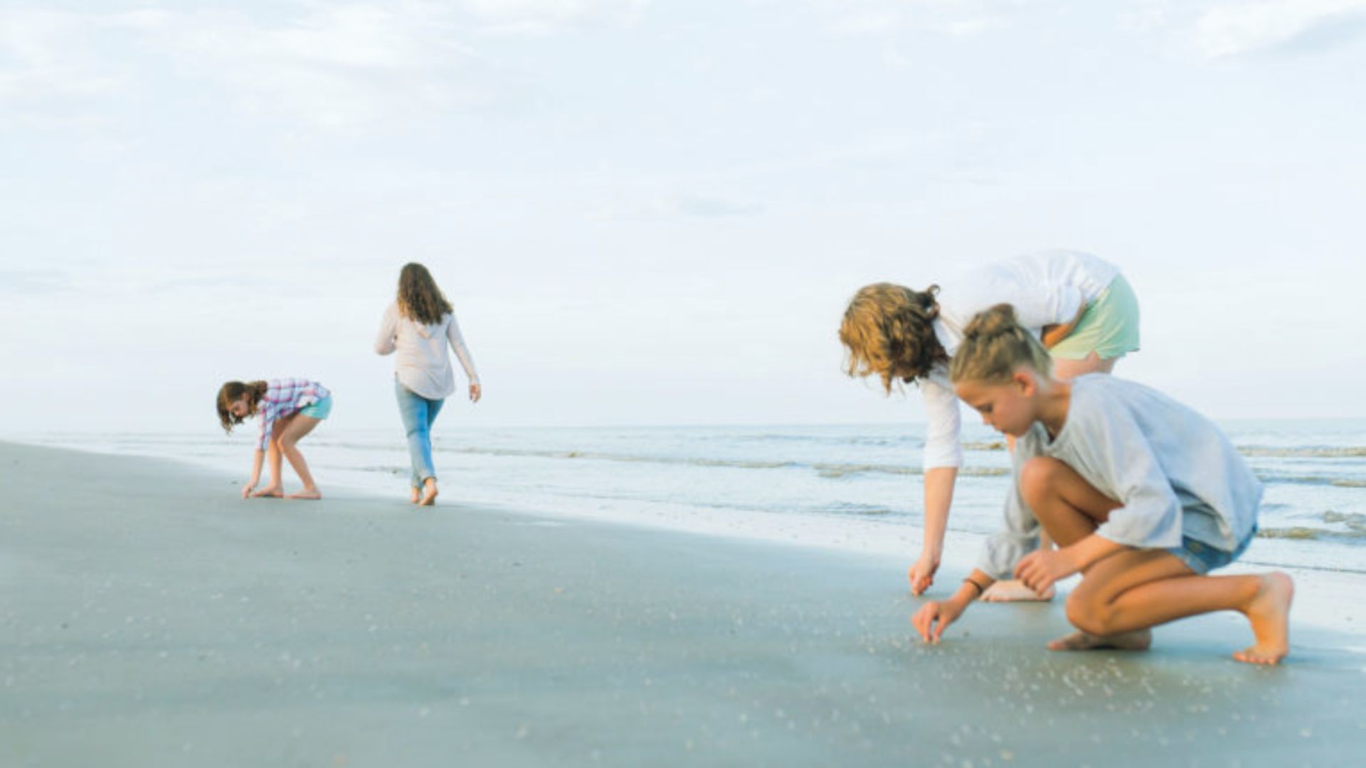 "Four children collect shells on an empty beach with calm ocean and cloudy sky."