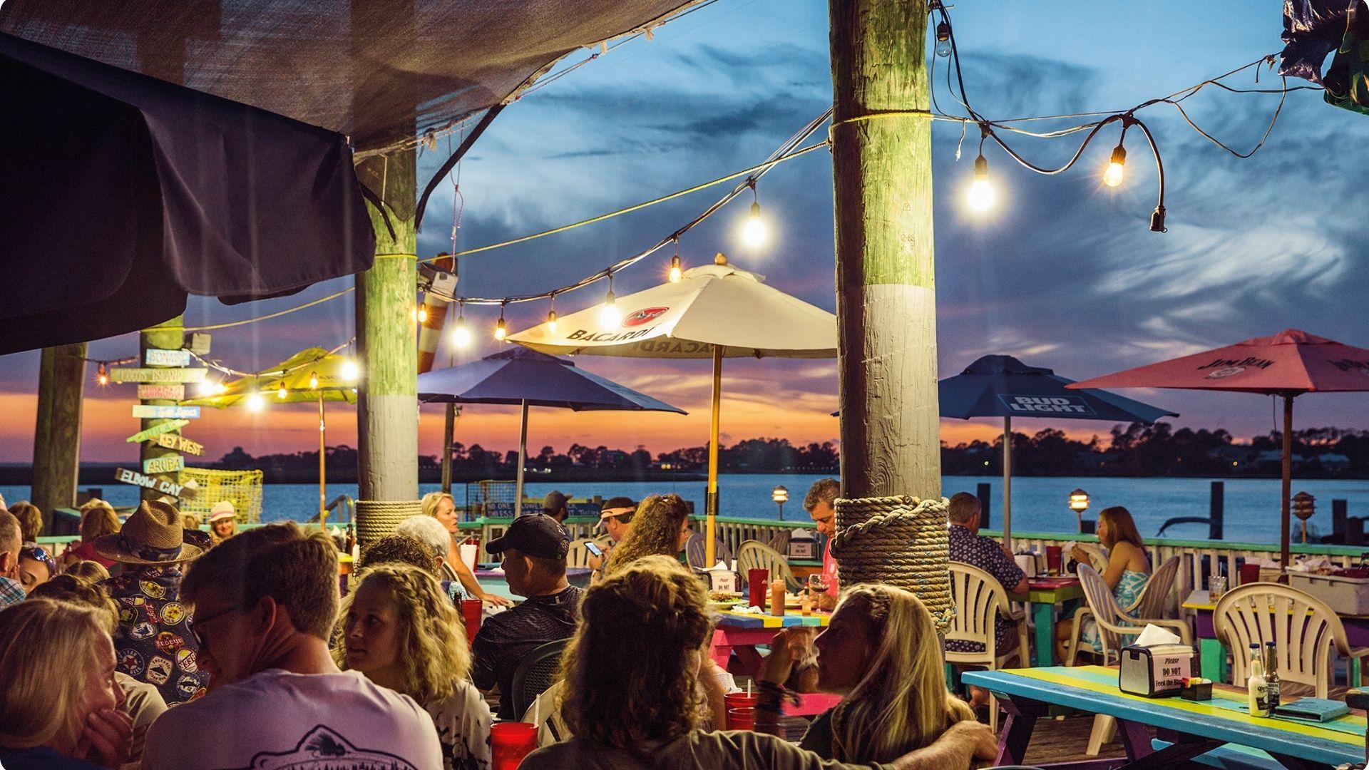 Vibrant outdoor waterfront dining scene at sunset with string lights, colorful picnic tables, Bud Light umbrellas, and a crowd enjoying drinks by the water.