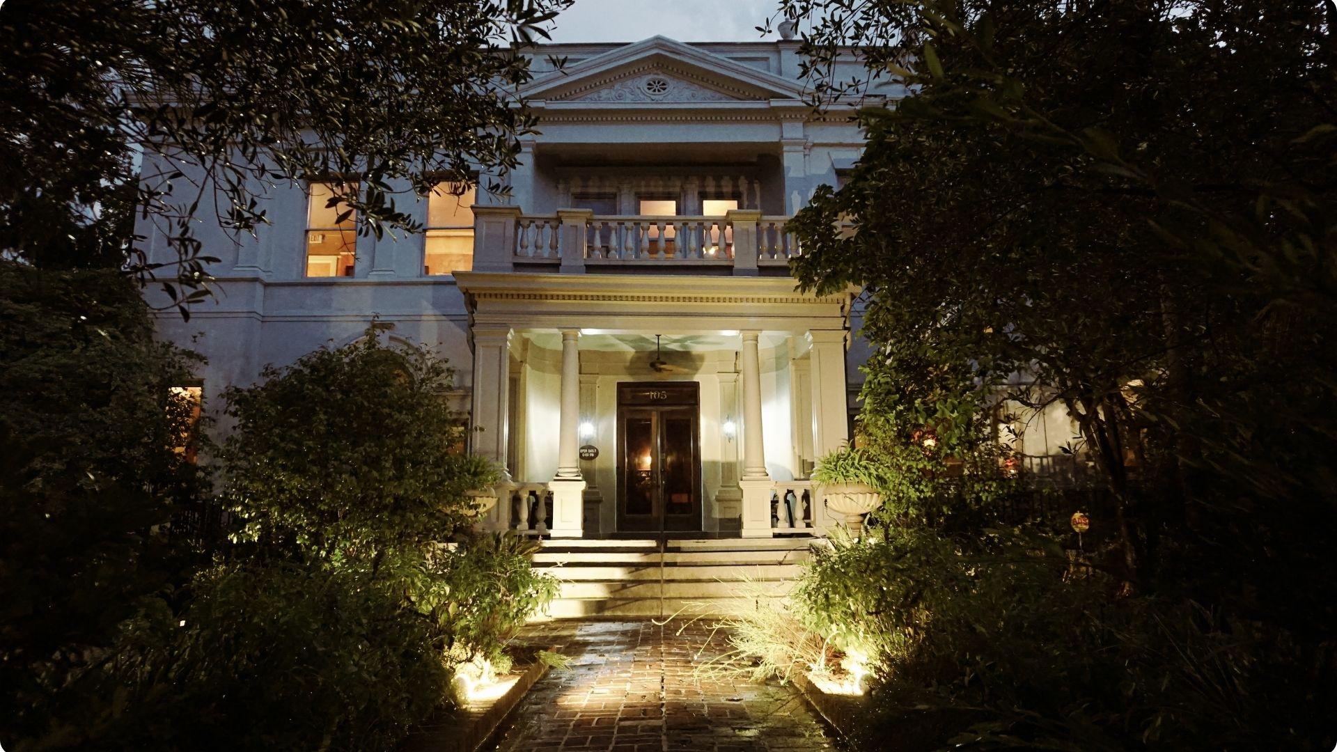 Dramatic nighttime exterior of a grand neoclassical white mansion with tall columns, balustraded balcony, and illuminated brick pathway framed by lush greenery.