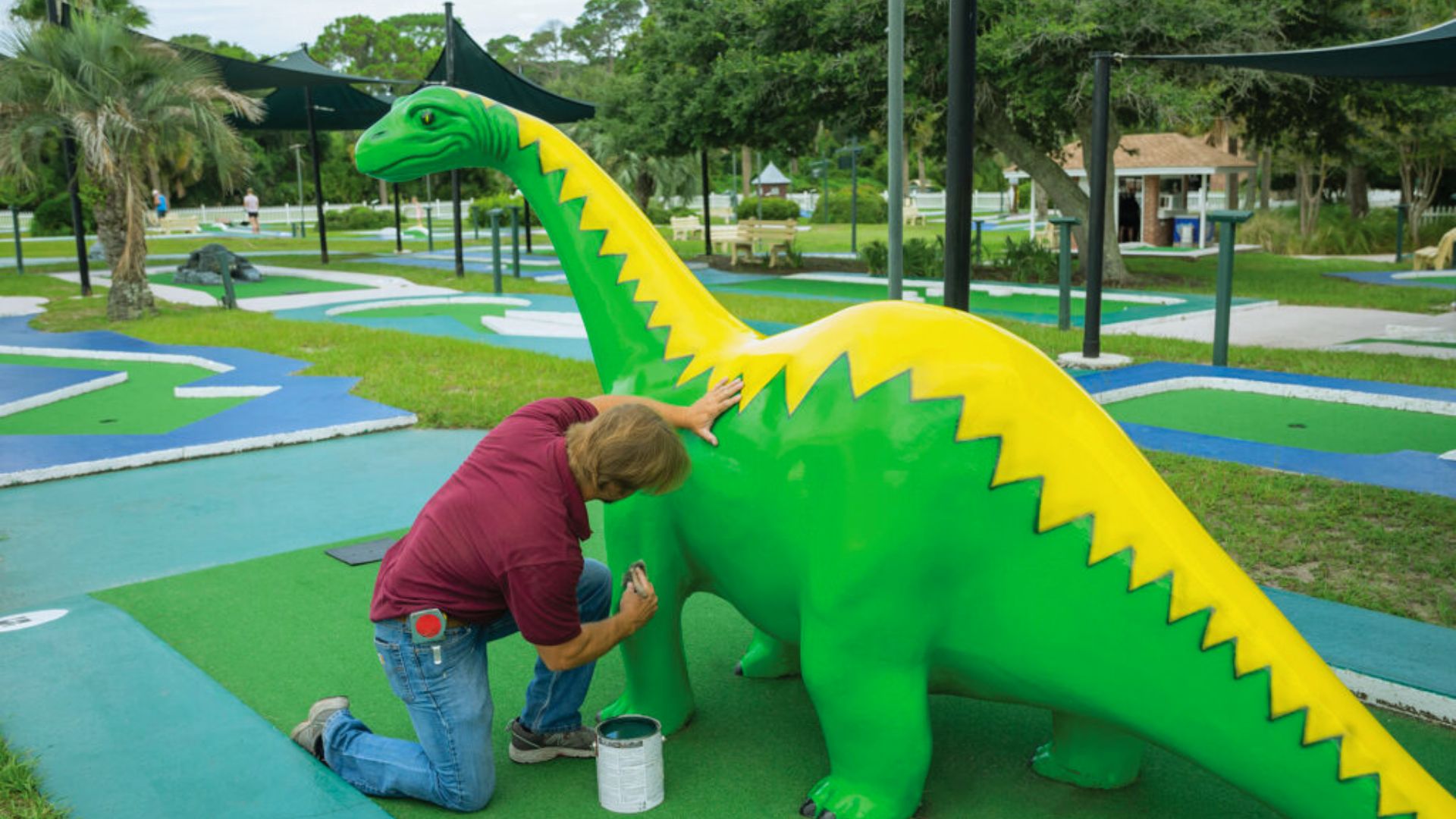 "Person kneels to paint a large green dinosaur statue on a mini-golf course."