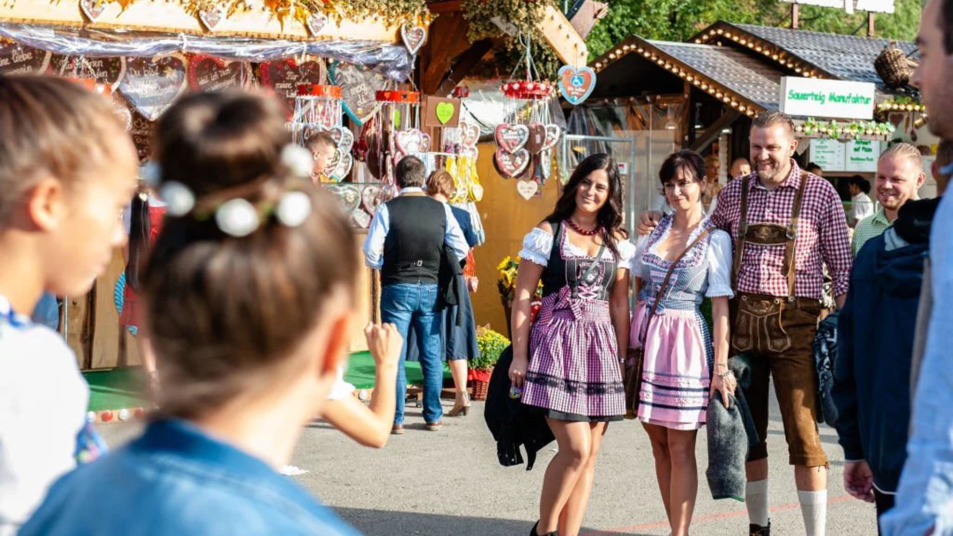 "People in dirndls and lederhosen smile at an outdoor festival with gingerbread heart decorations at market stalls."