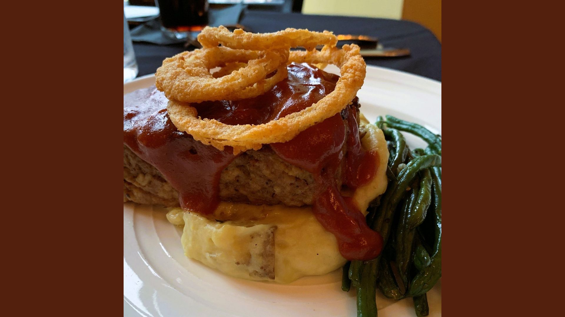 "Close-up of meatloaf topped with BBQ sauce and crispy onion rings, served over mashed potatoes with a side of sautéed green beans on a white plate"