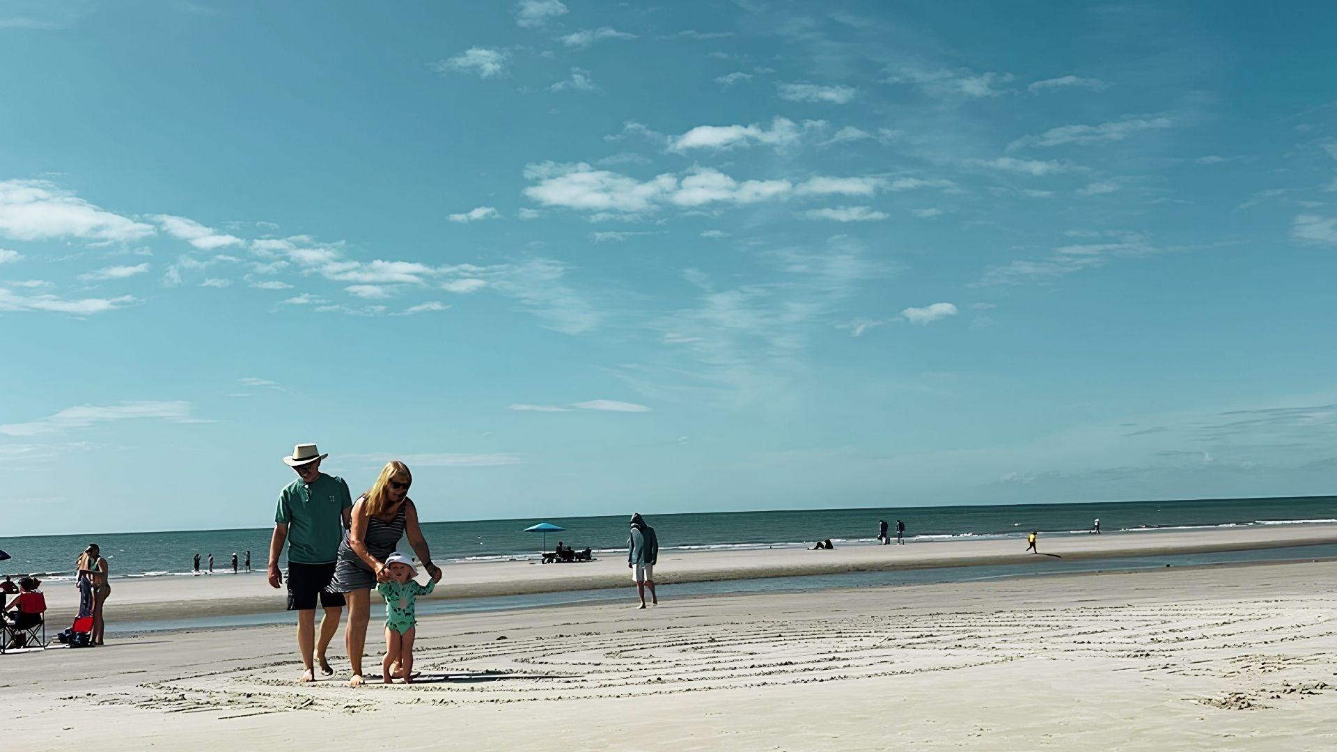 Family of three walking on sandy beach with toddler, ocean and beachgoers in background.