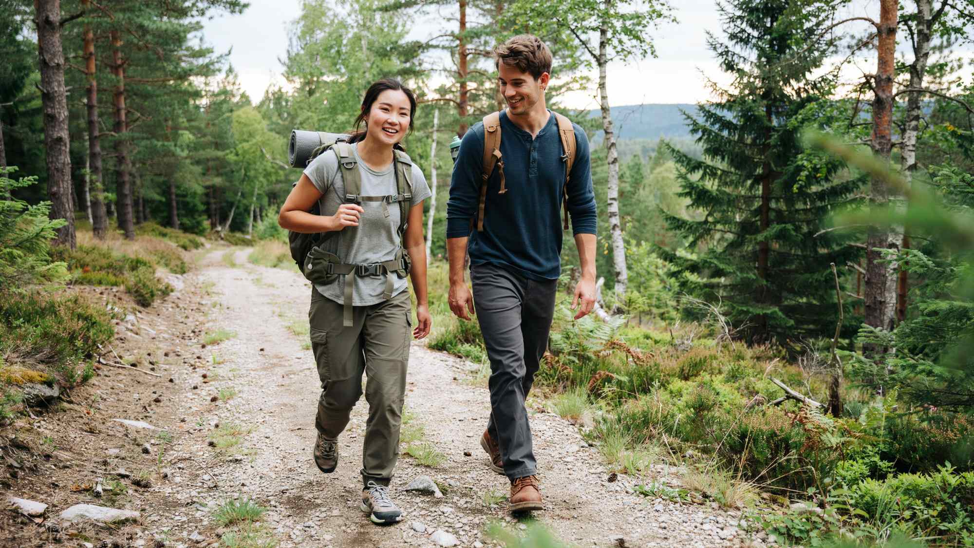 Two smiling hikers with backpacks walk together along a forest trail.