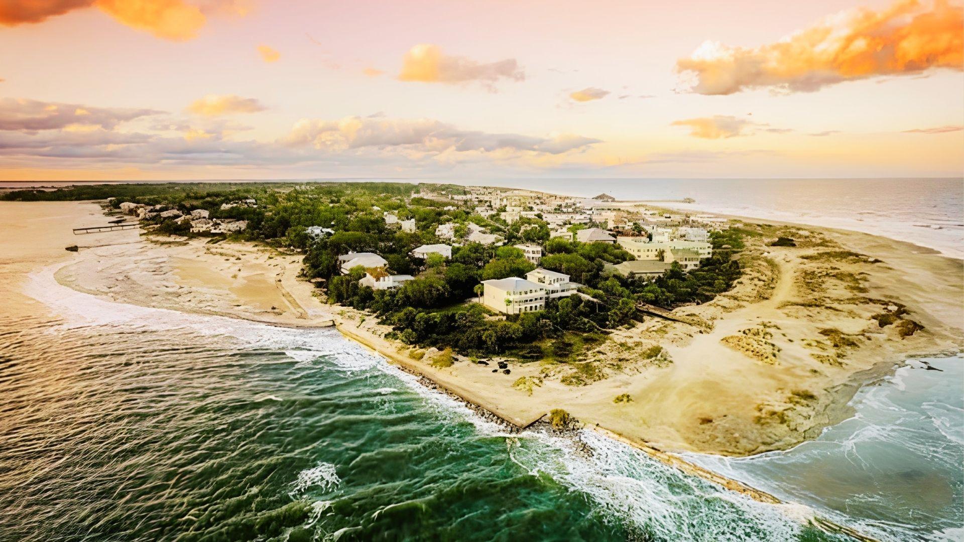 Aerial view of coastal island town with sandy beaches, houses among green trees at sunset.