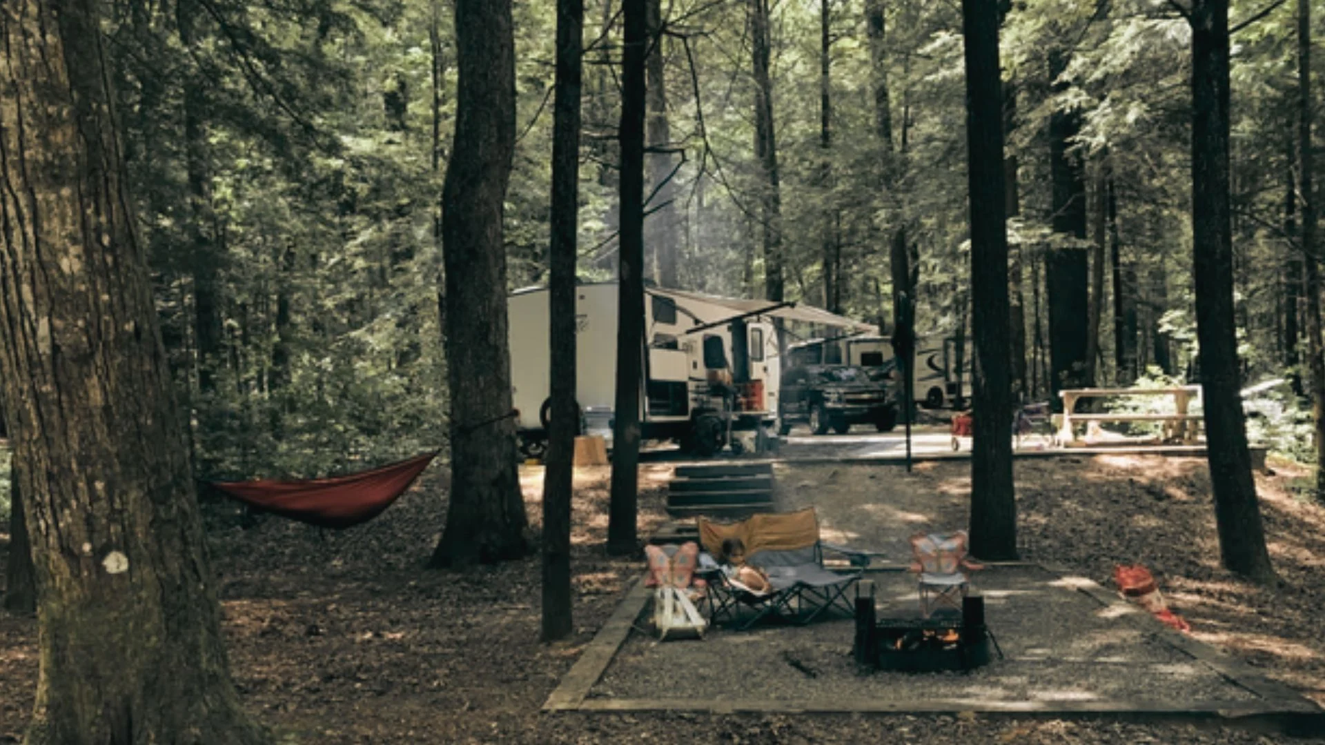 Travel trailer, hammock, and campfire area among tall trees at a shaded forest campsite setting.