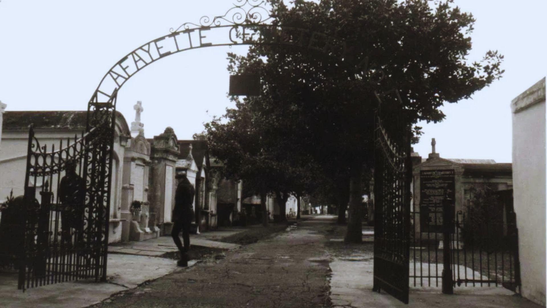 Sepia photograph of Lafayette Cemetery No. 1 entrance with iron archway, open gates, above-ground tombs, trees, visitor walking