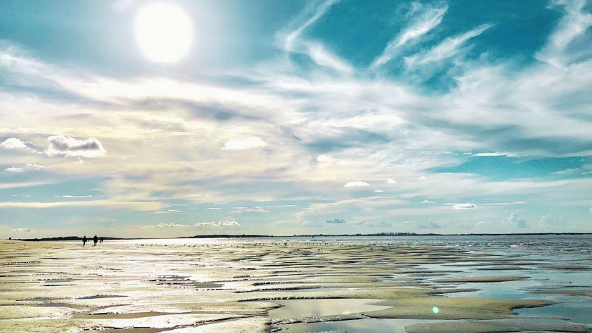 Sunny beach tidal flats with shallow pools reflecting sun, two silhouettes walking in distance.