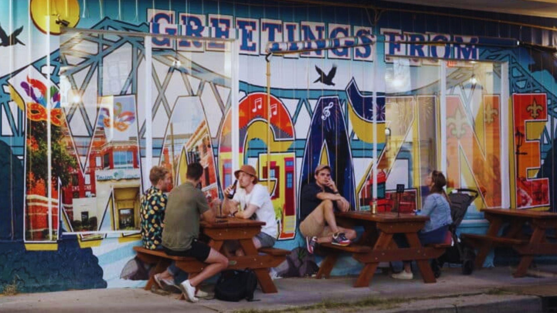 People seated at picnic tables before colorful "Greetings from Magazine" mural featuring bridge, music, fleur-de-lis, Mardi Gras