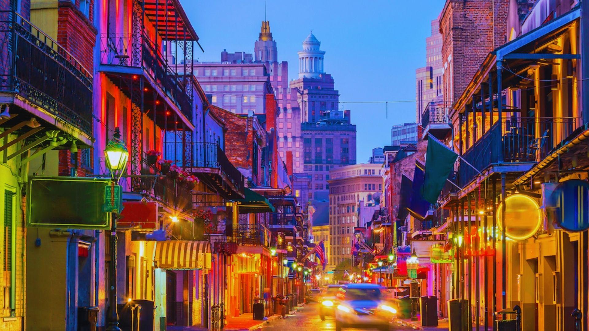 Bourbon Street at dusk glowing with neon lights, gas lamps, balconies, motion-blurred cars, French Quarter