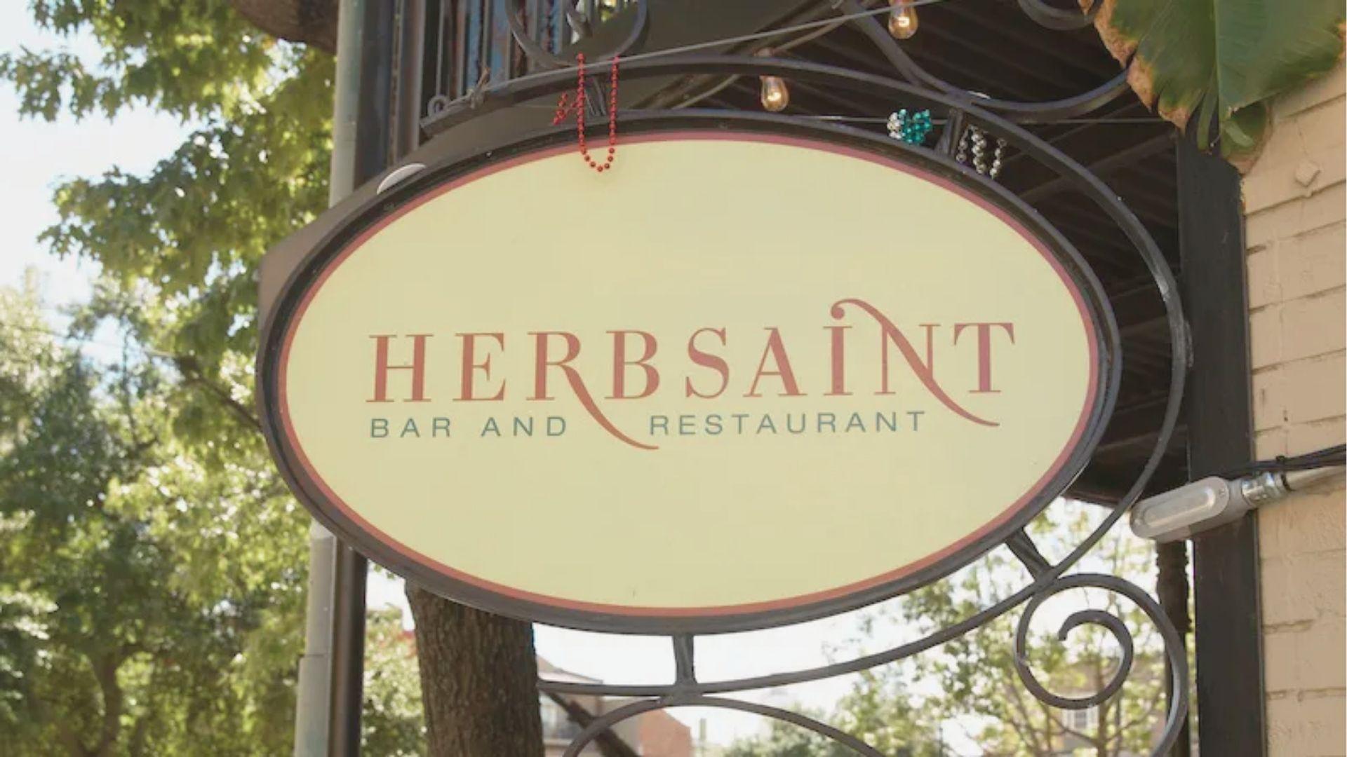 Oval cream-colored sign for Herbsaint Bar and Restaurant hanging from black ironwork, framed by green trees.