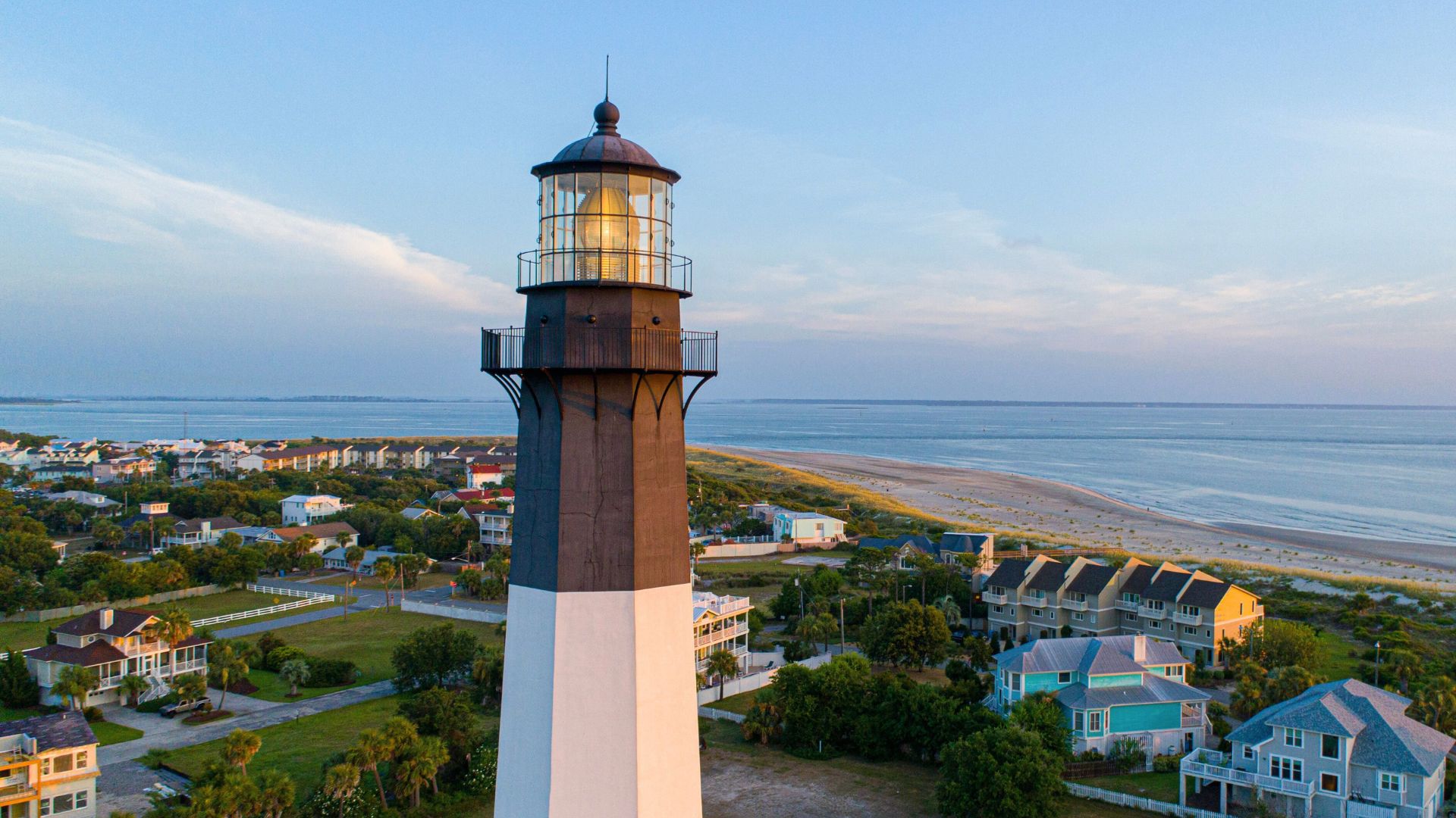 Tybee Island Lighthouse rises above beach houses and sandy shoreline beside calm ocean waters at sunset.
