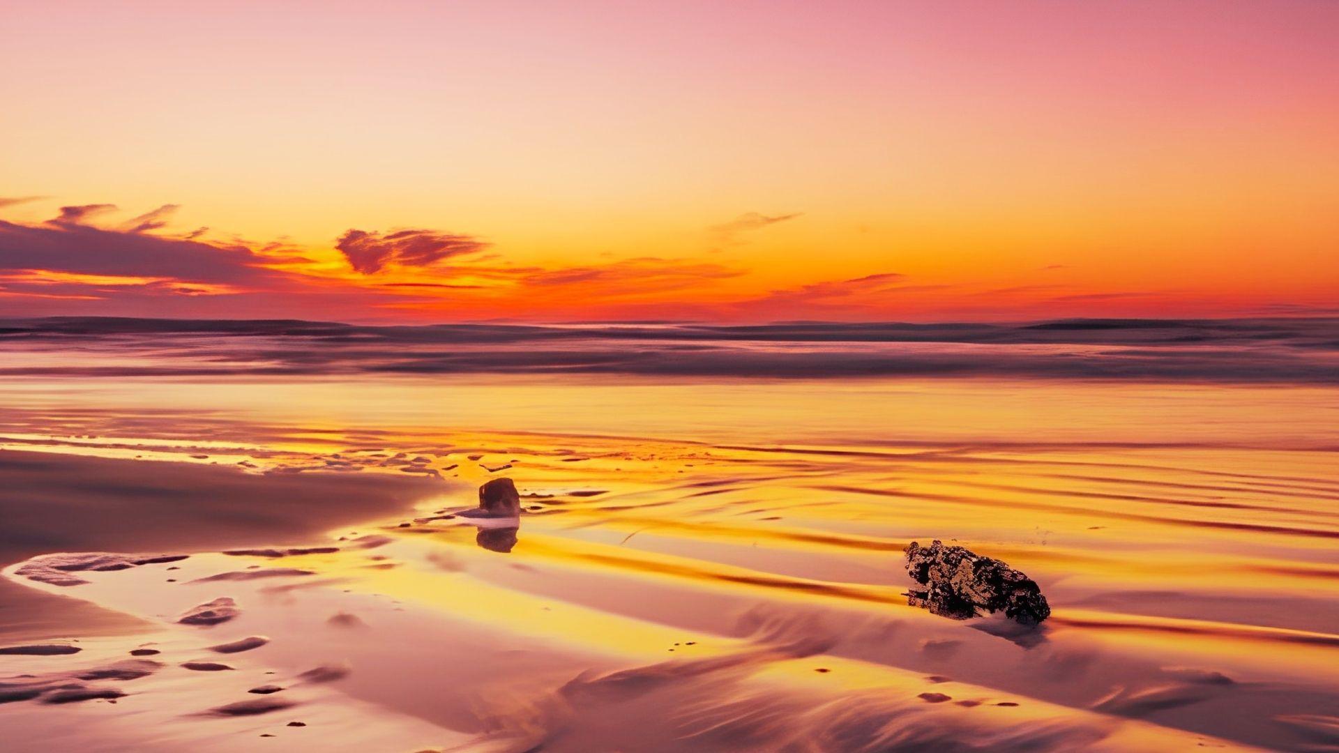 Golden orange sunset over wet sandy beach reflecting sky with rocks in shallow water.