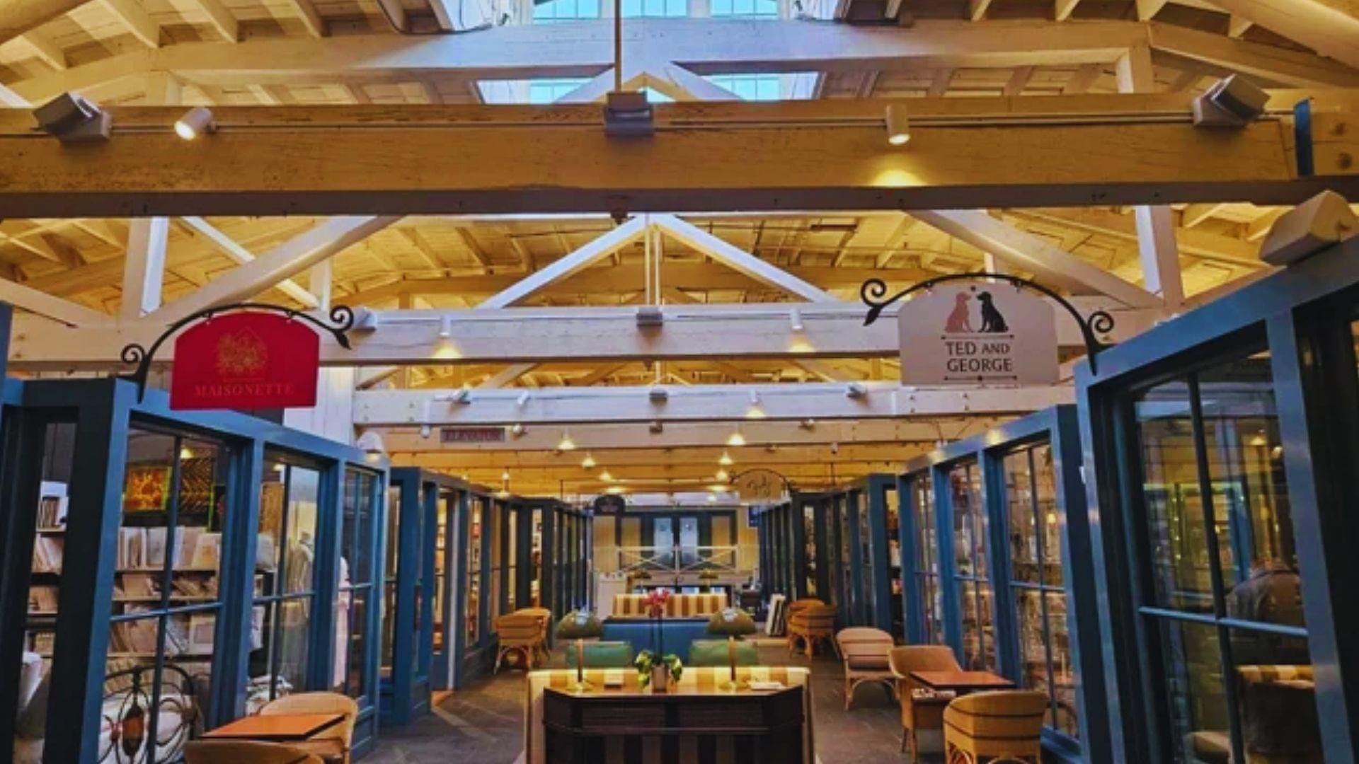 Sunlit indoor shopping arcade with blue storefronts, wooden beams, signs for Maisonette and Ted and George boutiques