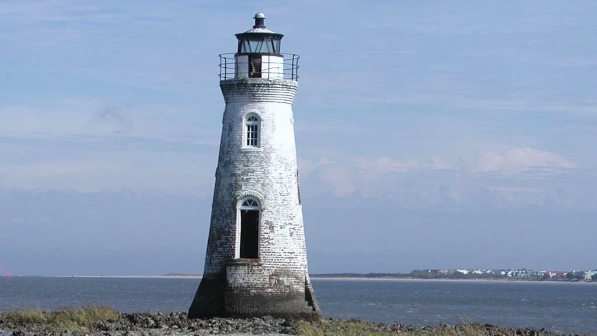 Cockspur Island Lighthouse rises from a rocky shore under a clear blue sky.