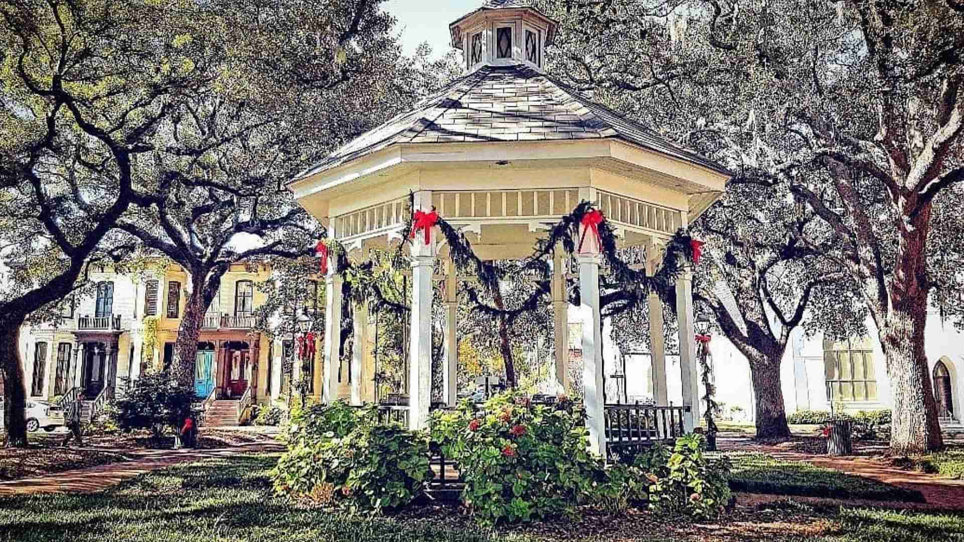 White gazebo decorated with Christmas garlands and red bows in Savannah square, framed by oak trees and historic homes