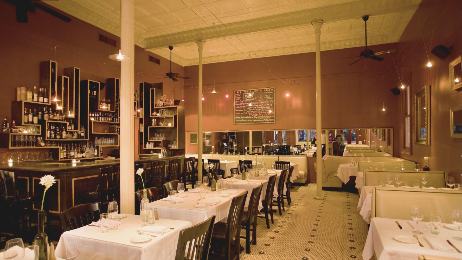 Classic New Orleans dining room with white-clothed tables, dark chairs, and a bar under warm pendant lights.