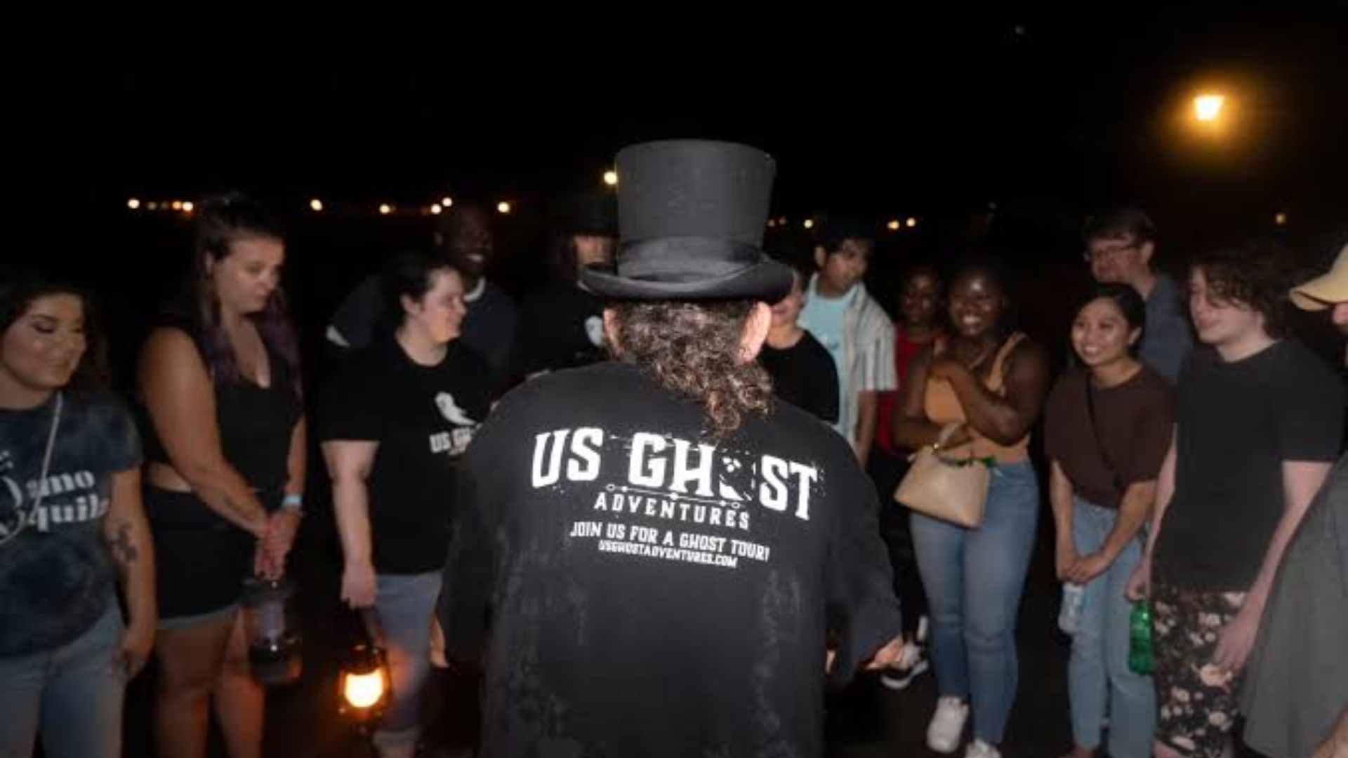 Tour guide in top hat and US Ghost Adventures shirt leading a circle of tourists at night holding lanterns.