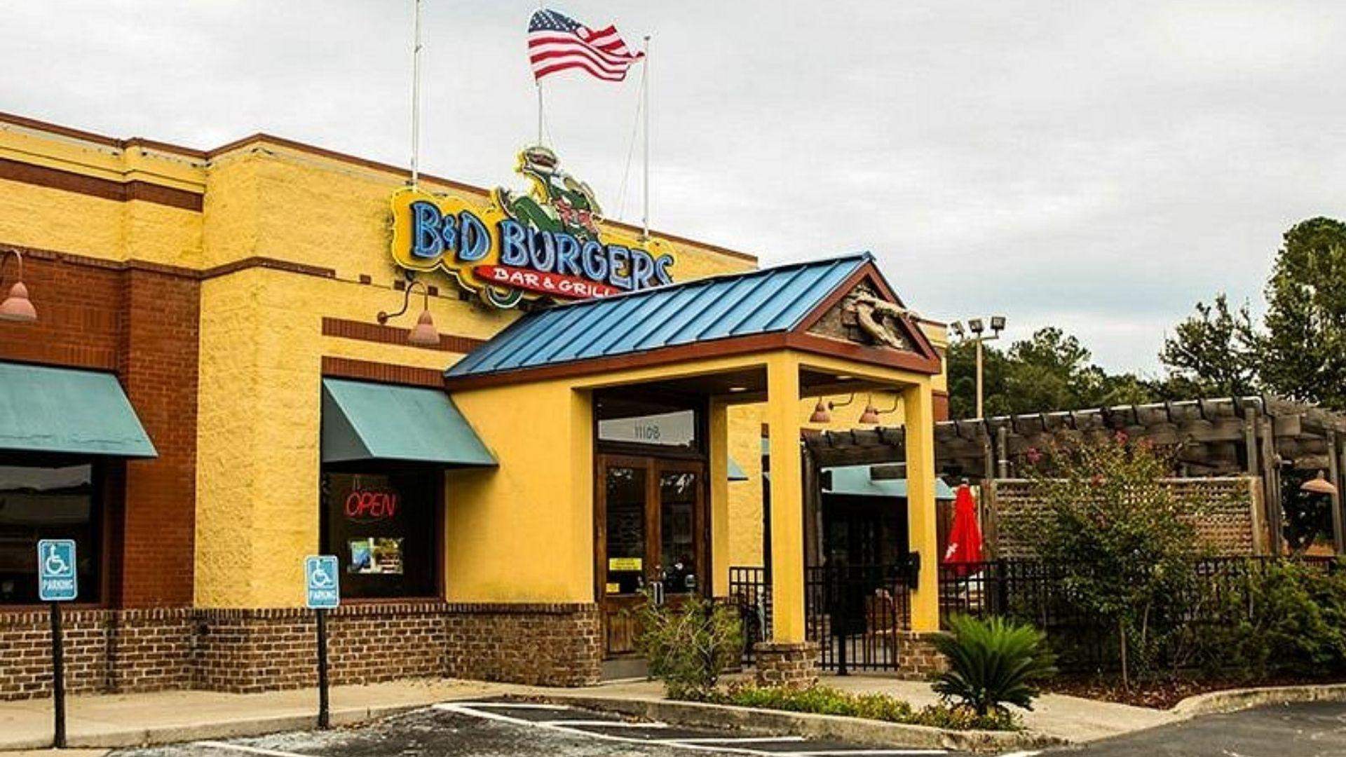 Yellow and blue exterior of B&D Burgers with hut shaped entrance, American flag on roof.